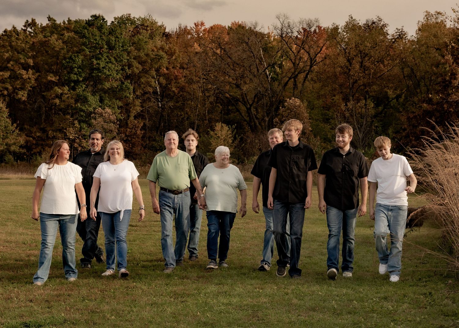 A large family group of nine people walking together across a green lawn with colorful autumn trees in the background.