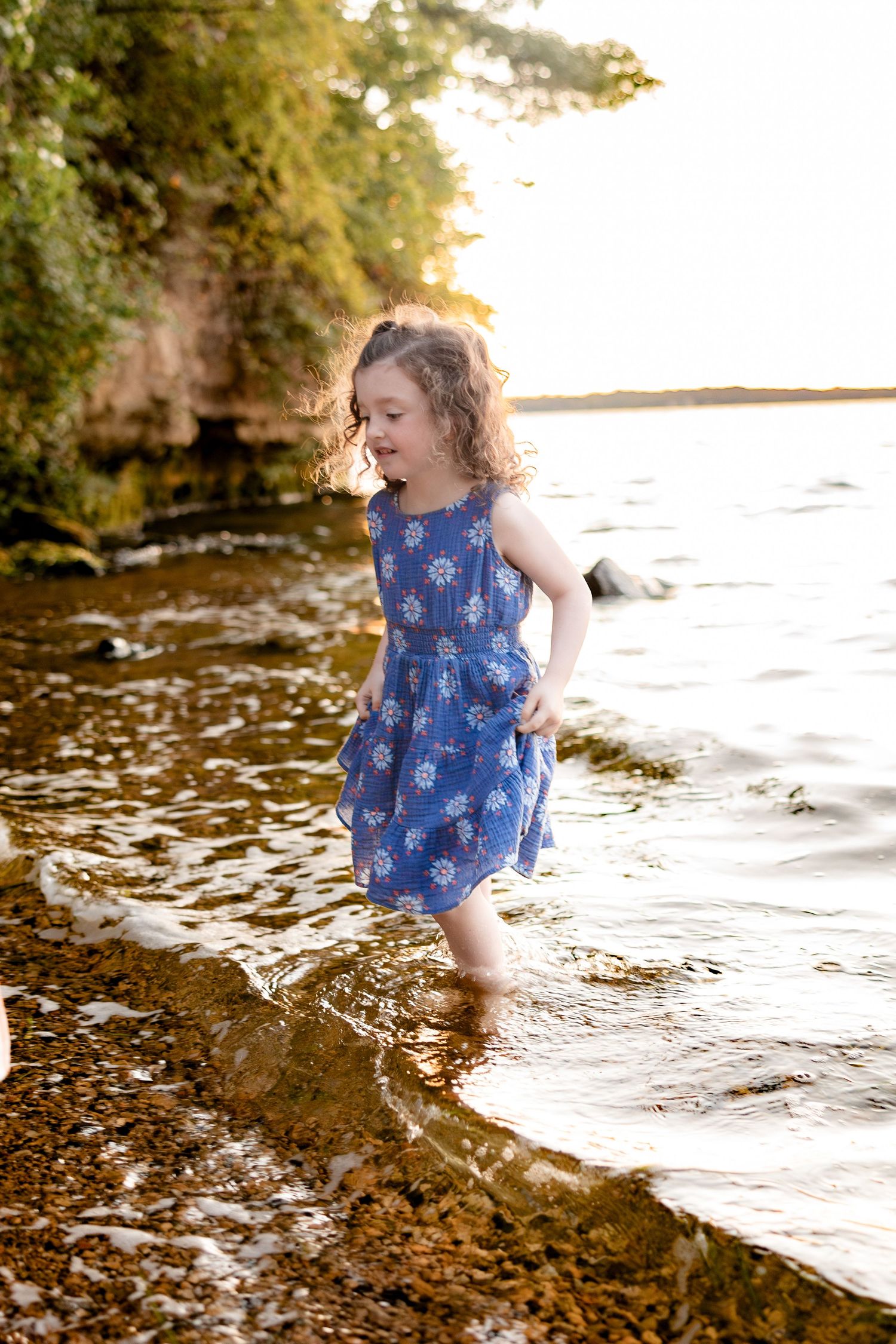 A young girl in a blue floral dress wades barefoot in shallow lake water at golden hour.