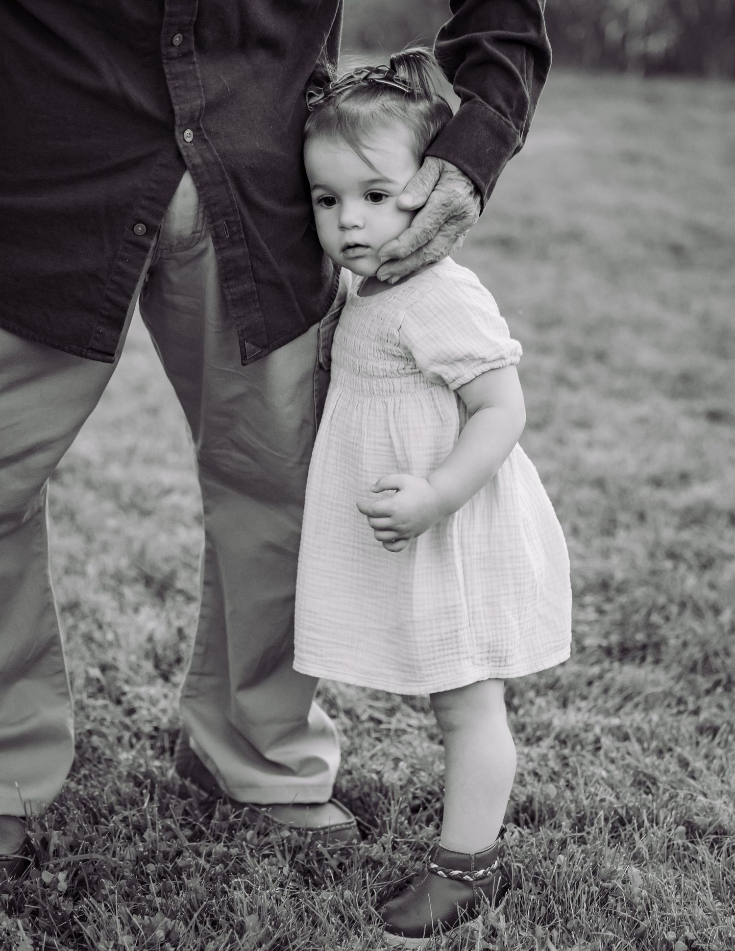 Young toddler girl in white dress standing beside adult holding a stuffed animal, black and white photo.