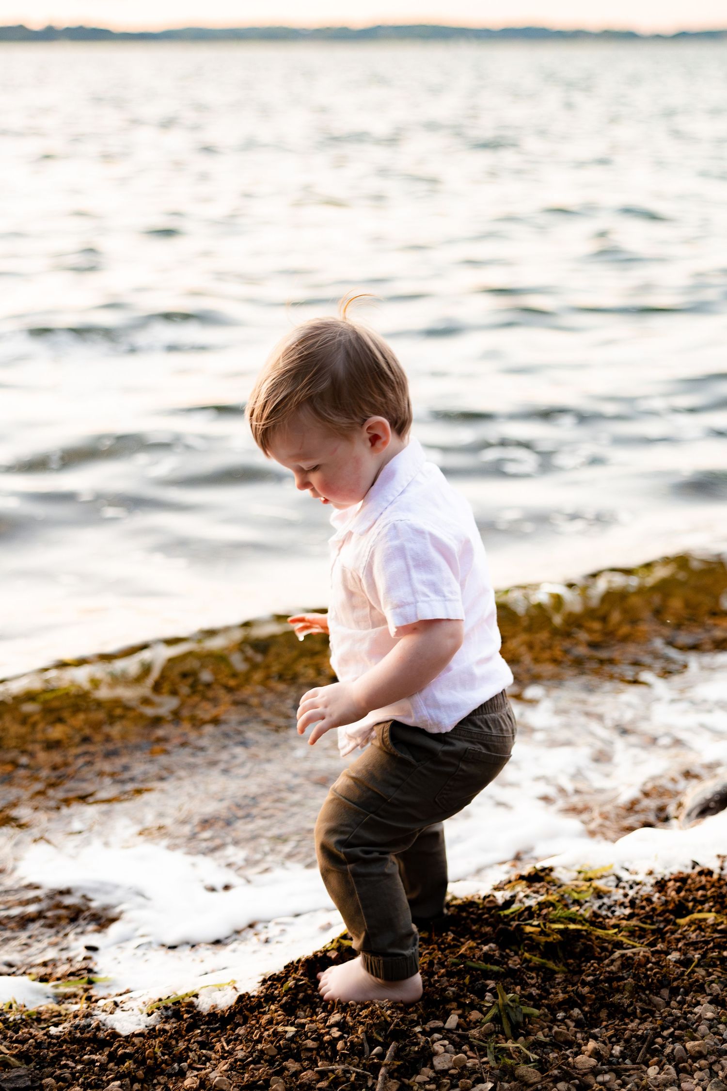 Toddler boy in white shirt and dark pants playing barefoot on rocky shoreline near gentle ocean waves at sunset.