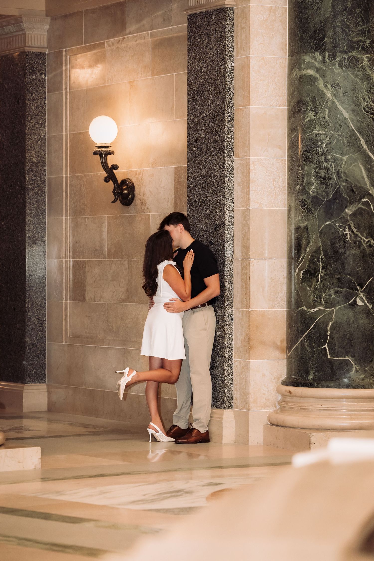 Couple sharing a romantic kiss in an elegant marble hallway, woman in white dress standing on tiptoes.