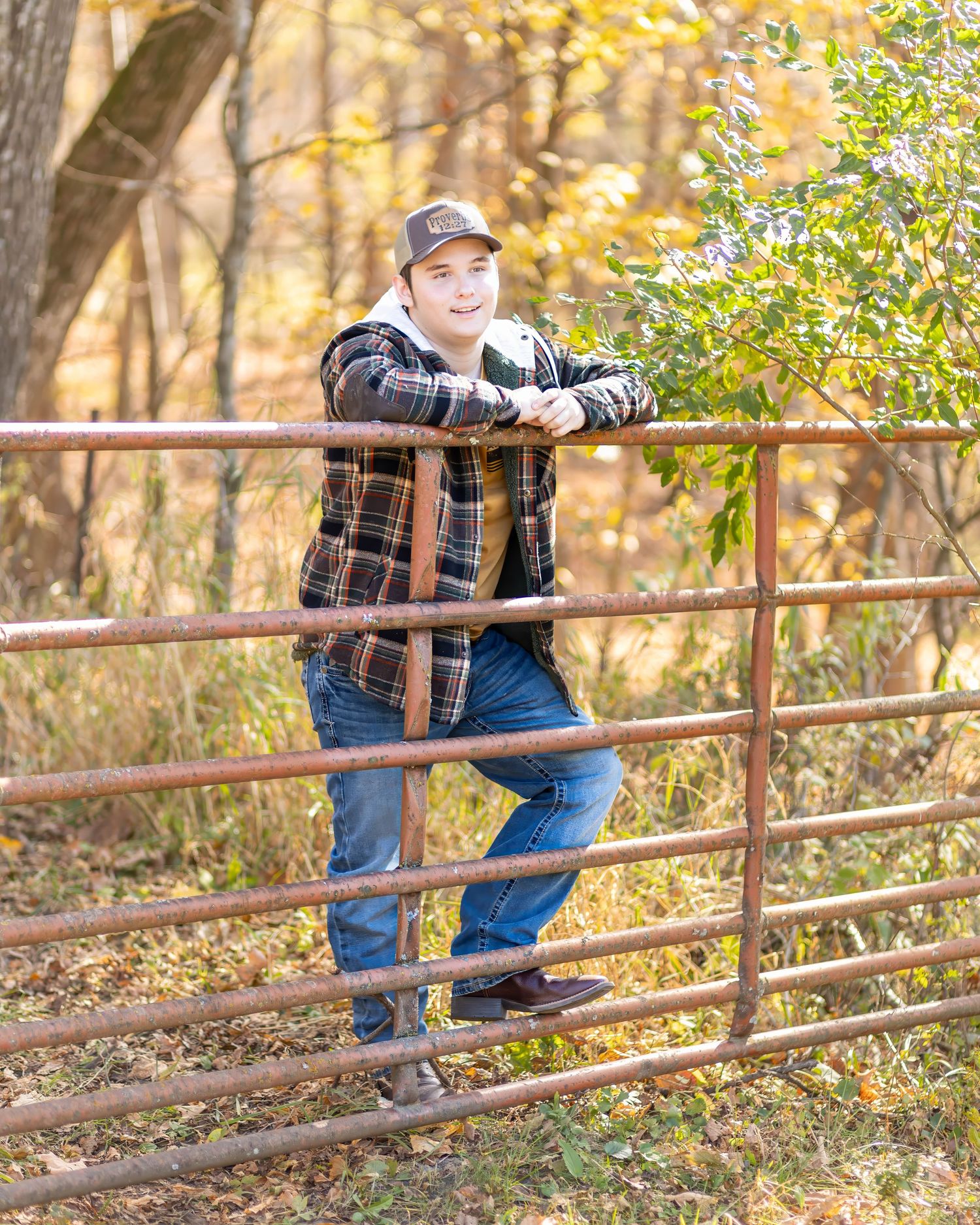 Smiling teen boy in flannel jacket leans on rustic metal gate among fall foliage senior portrait.