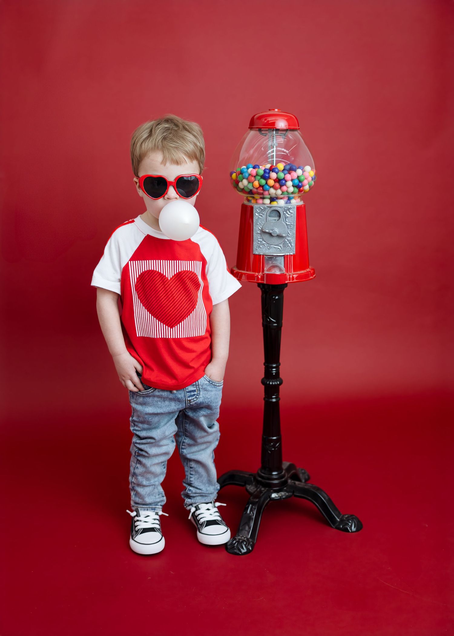 Cool toddler blows bubble gum beside gumball machine on red background.