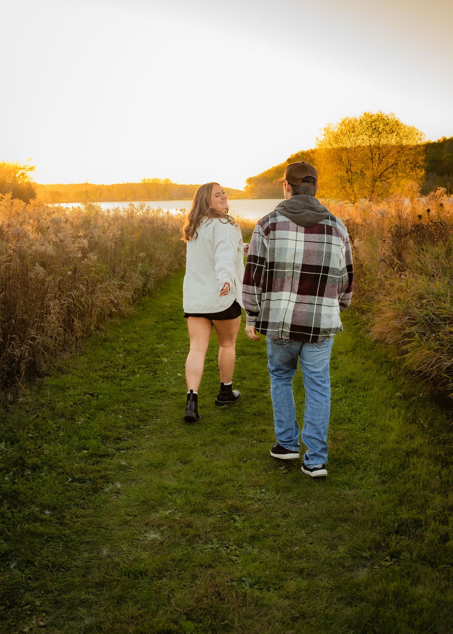 Couple walking hand in hand along grassy trail toward glowing sunset over tranquil lake.