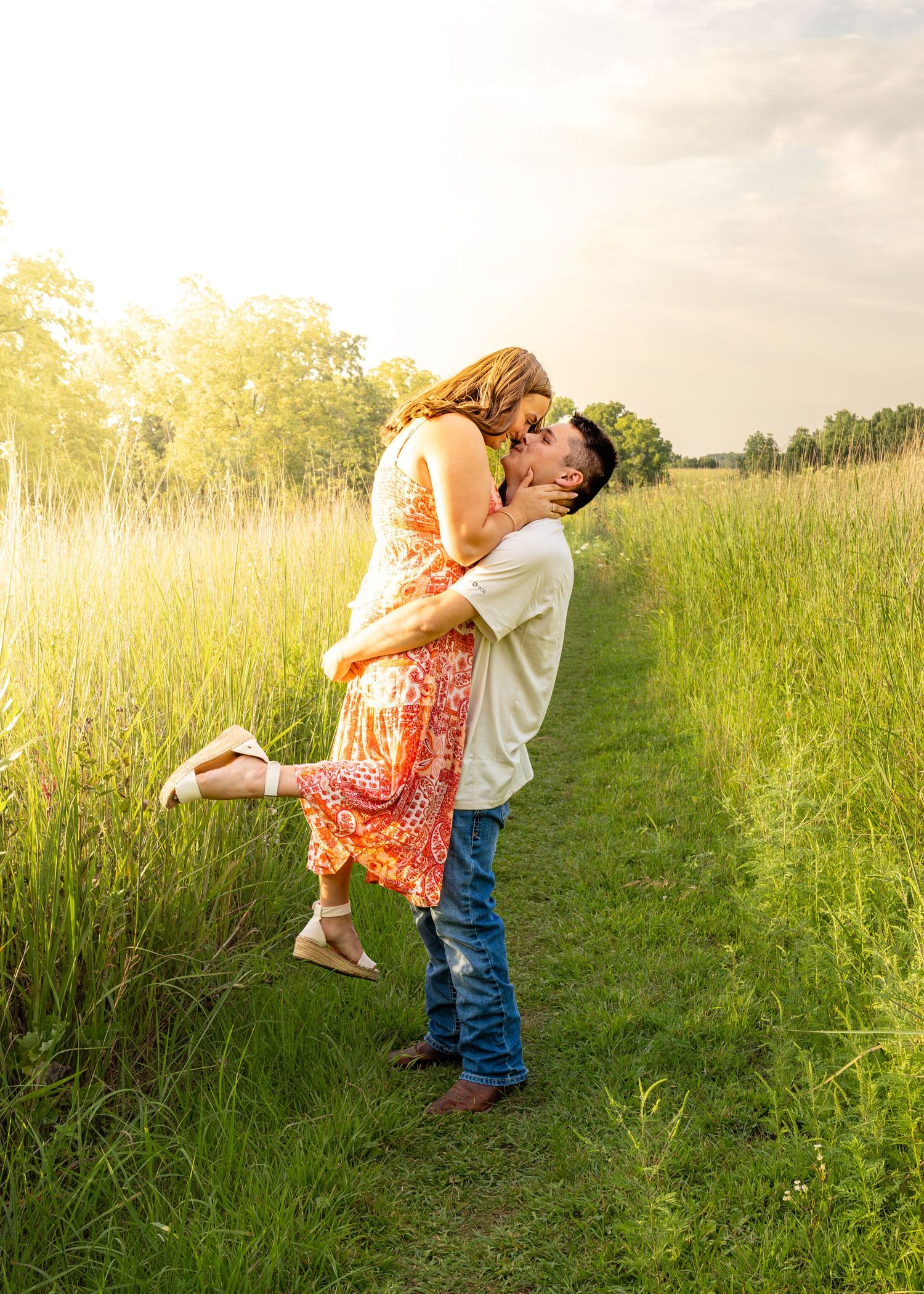 Man lifting woman in red floral dress for a kiss along a lush green field path at golden hour.