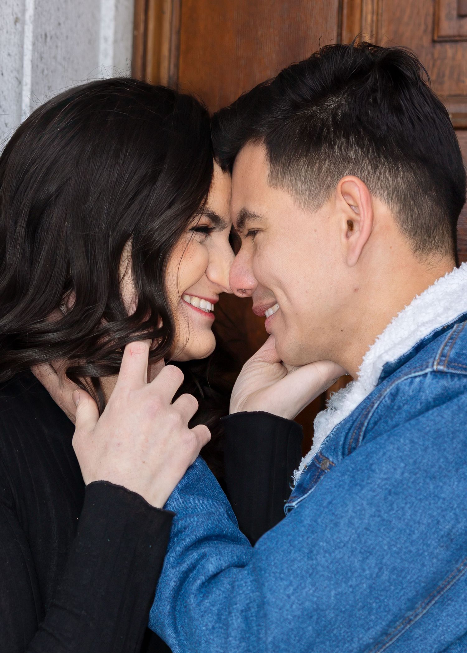 Smiling couple leans close together in front of a wooden door, woman gently touching mans chin lovingly.