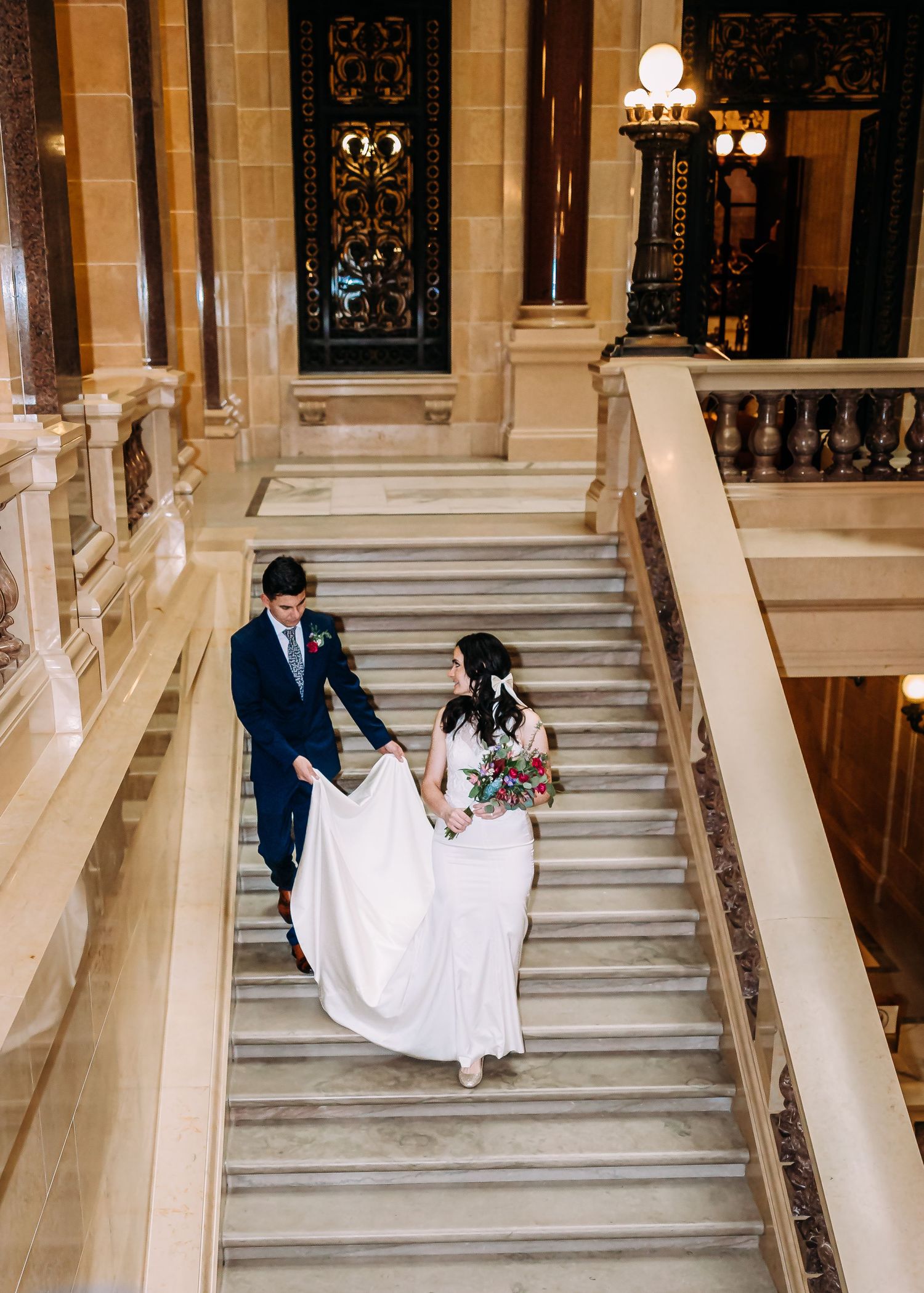 Aerial view of bride in white gown and groom in navy suit walking down grand marble staircase.