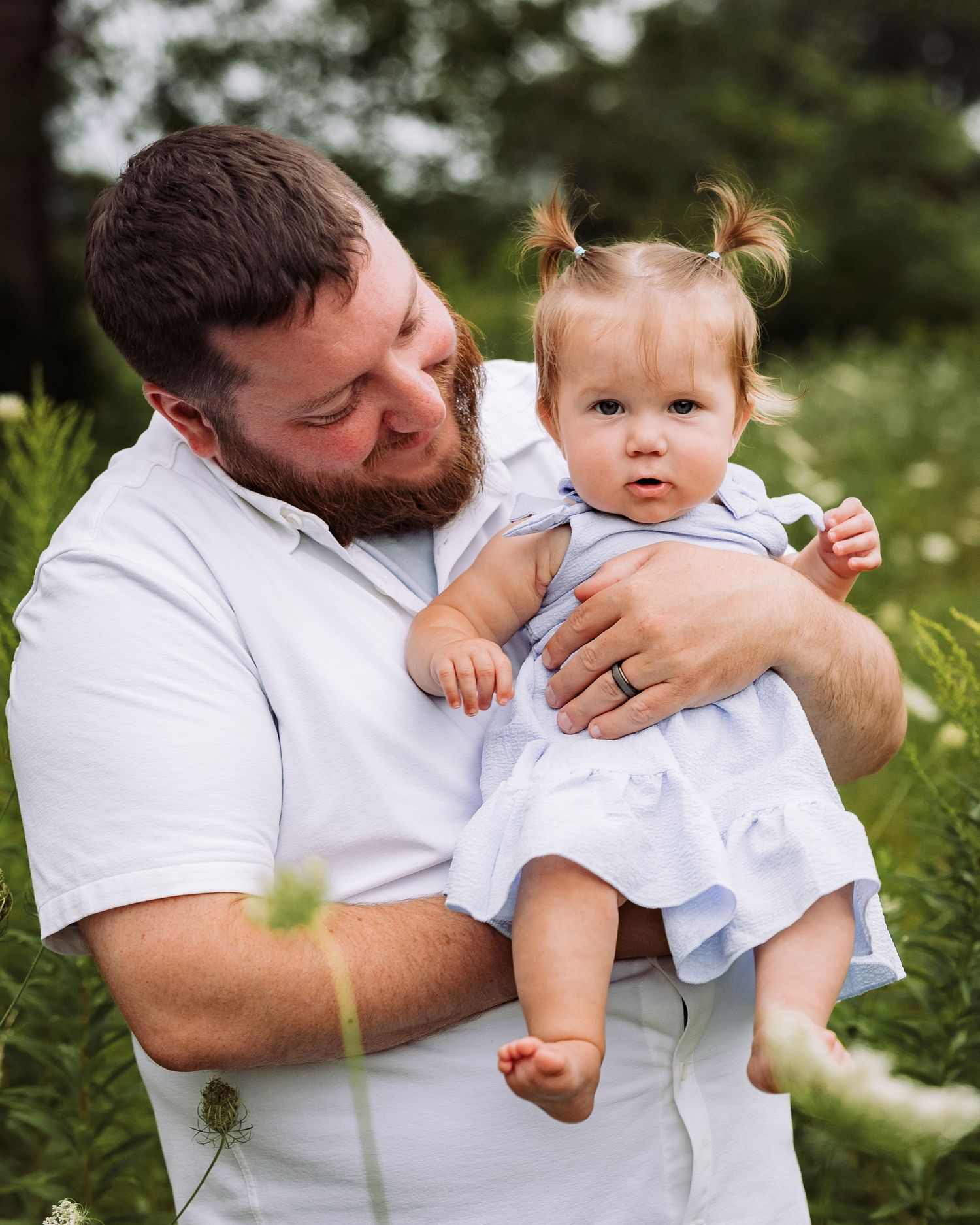 A bearded man in a white shirt holds a baby girl in a blue dress outdoors in a lush green field.