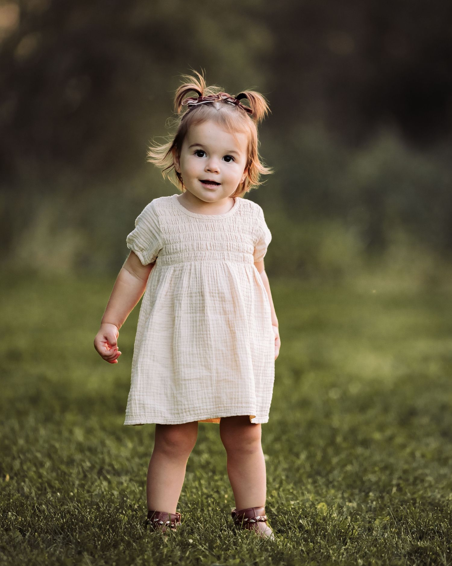 Adorable toddler girl in a white smocked dress standing on green grass outdoors in soft natural light.