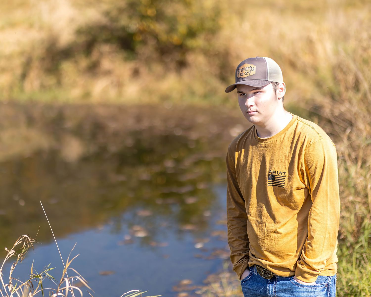 Young man in mustard yellow long-sleeve shirt and cap stands near a pond in a golden autumn field.