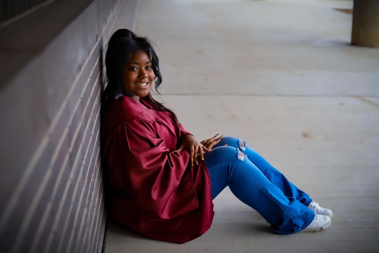 A young woman in a maroon graduation gown sits smiling against a wall in ripped jeans.