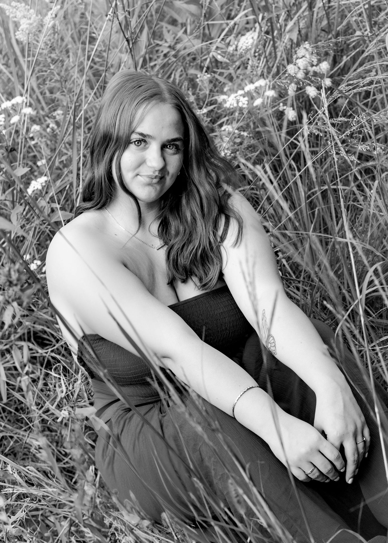 Black and white senior portrait of young woman sitting in tall grass wearing strapless dress.