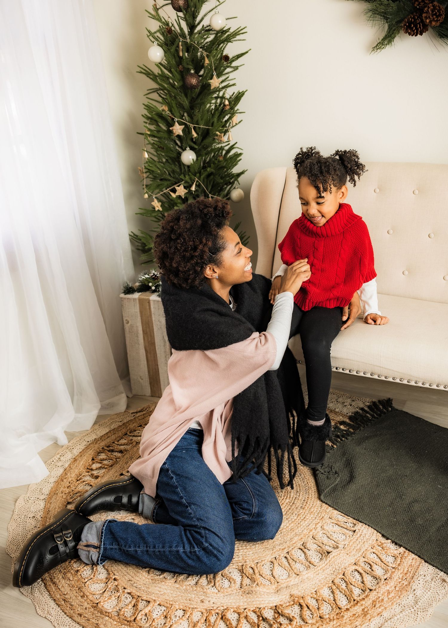 A mother kneels on a rug helping her young daughter in a red sweater near a decorated Christmas tree.