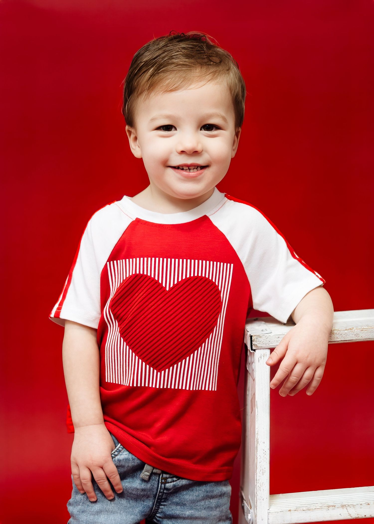 Happy toddler in red and white heart shirt smiles beside white ladder.