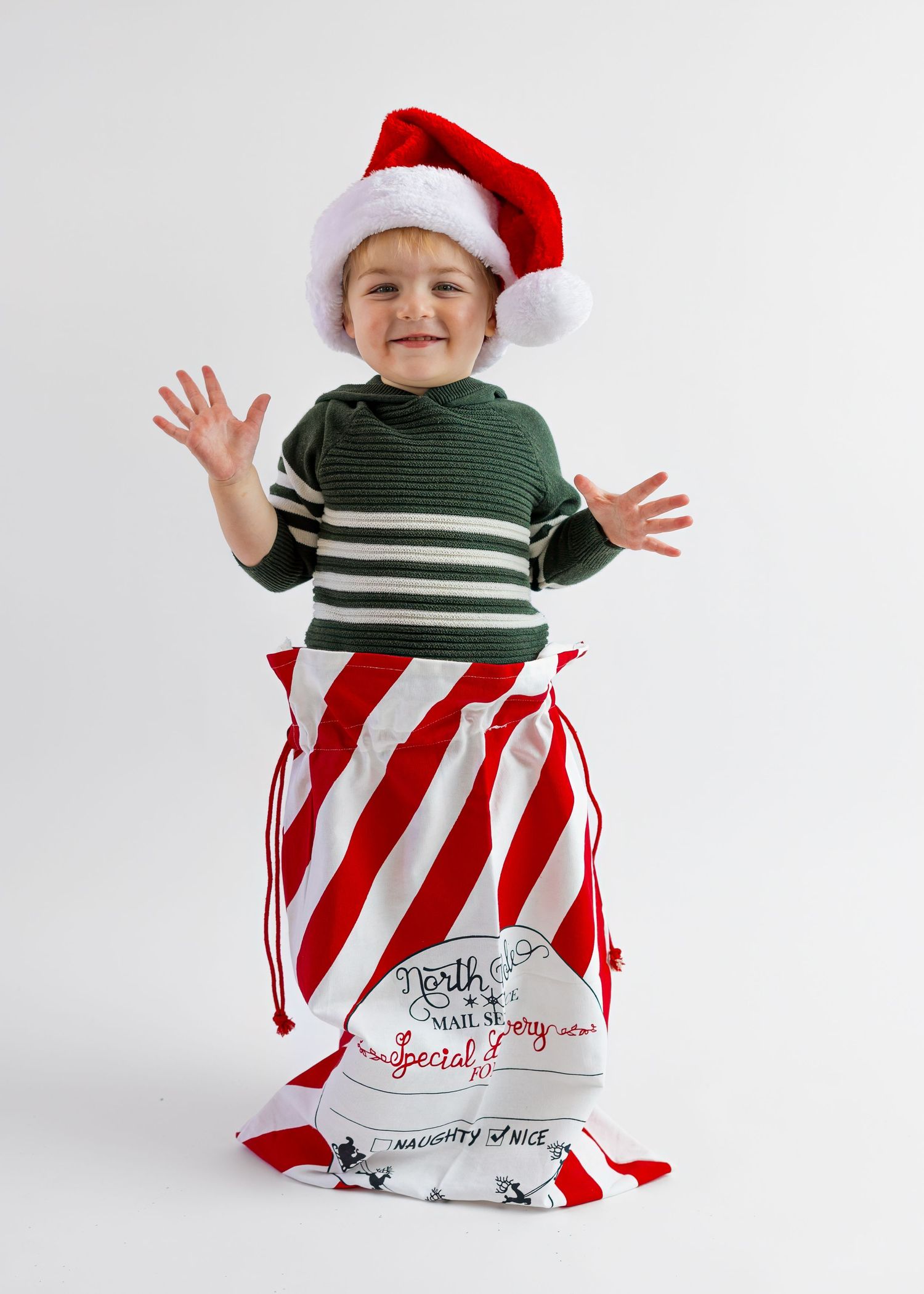 Joyful toddler in striped sweater and Santa hat raises hands inside a red striped Christmas sack.