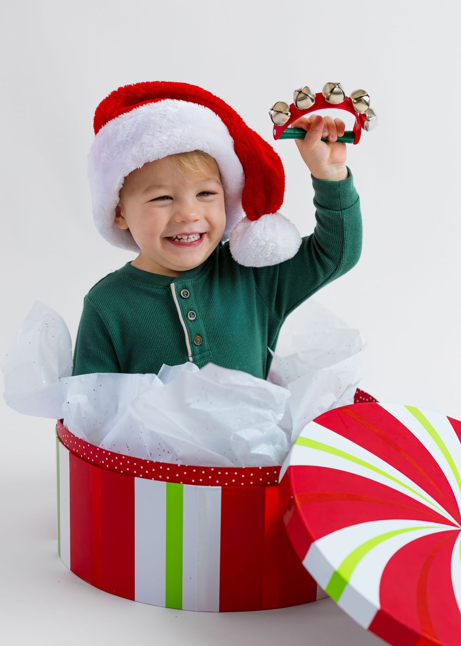 Joyful toddler in Santa hat popping out of festive gift box holding jingle bells at Christmas.