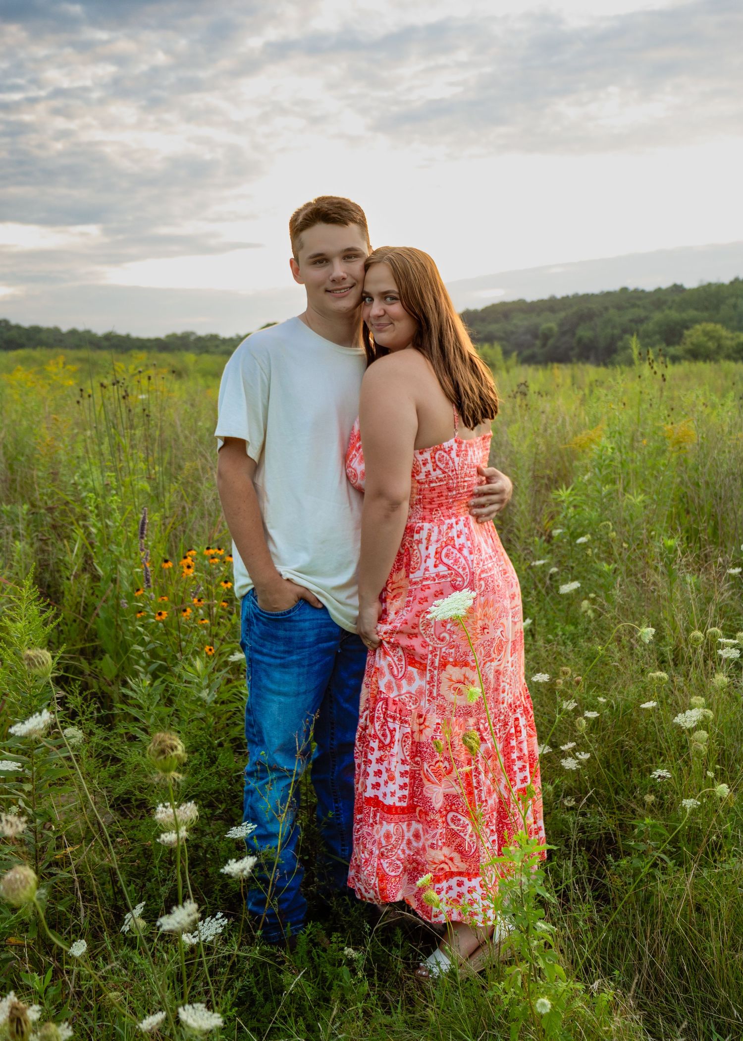 Young couple embracing in a wildflower meadow at sunset, woman in pink floral maxi dress.