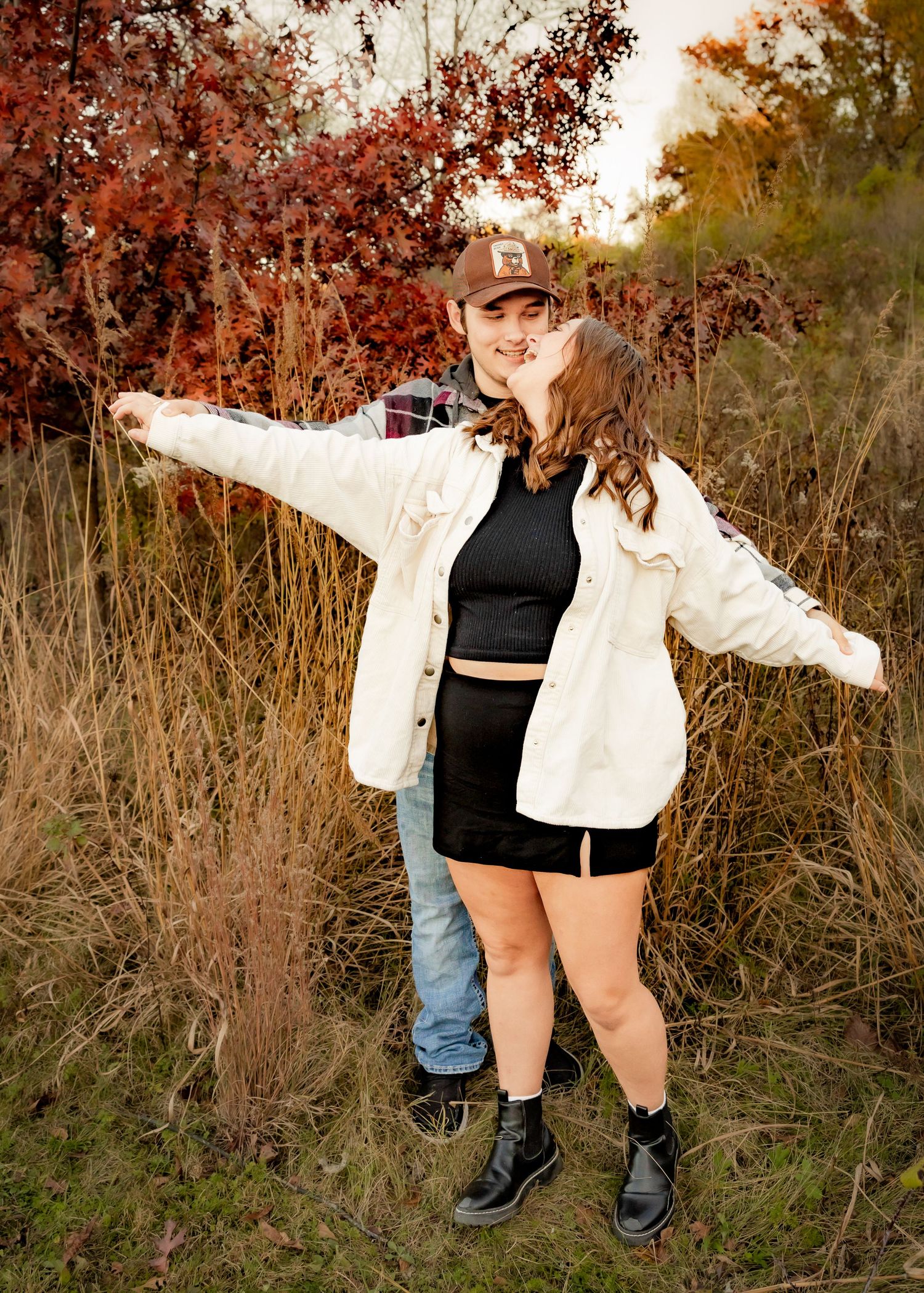 Couple posing playfully in tall grass with vibrant red fall foliage in background.