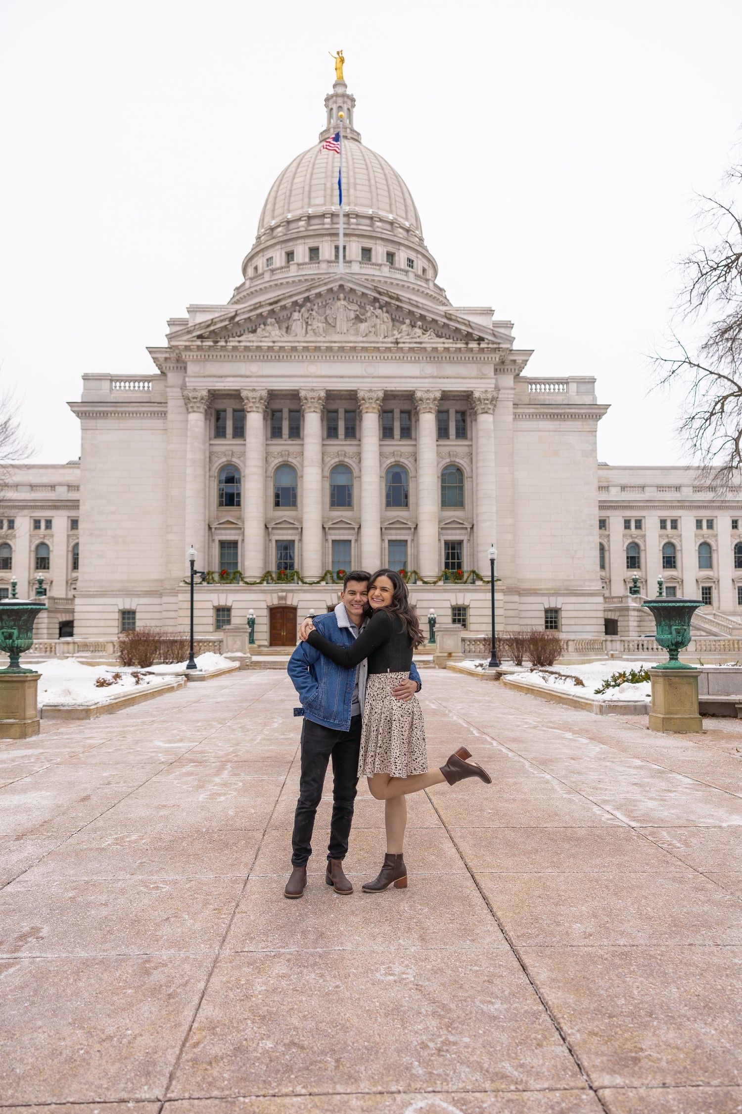 A happy couple embraces in front of a grand domed capitol building surrounded by snow-covered grounds.
