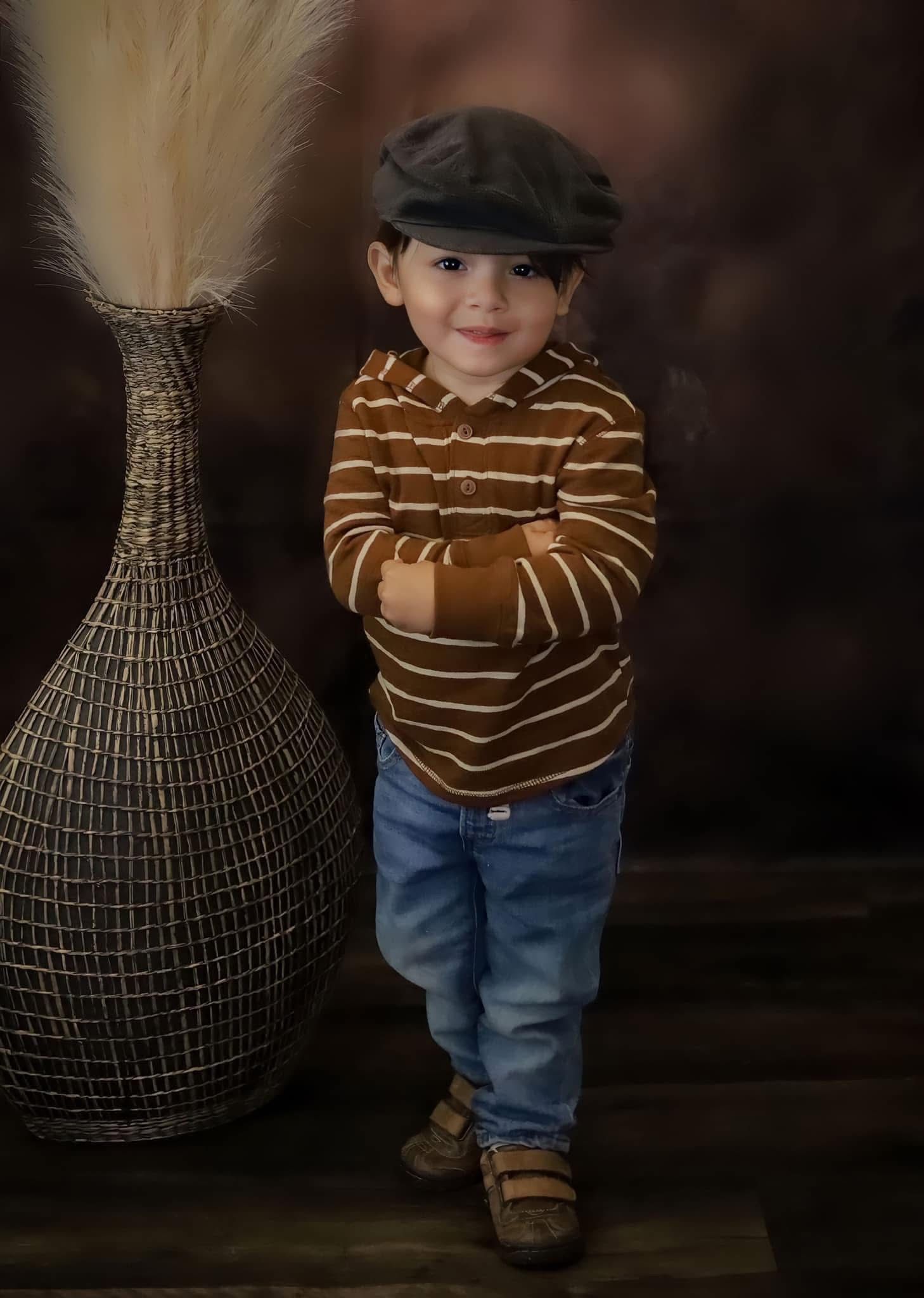 Smiling toddler boy in striped brown shirt and newsboy cap stands confidently with arms crossed.