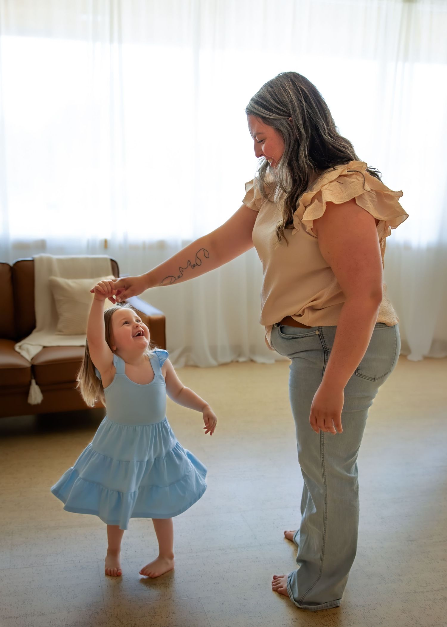Mother and daughter dancing joyfully indoors, girl in blue dress spinning in bright living room.