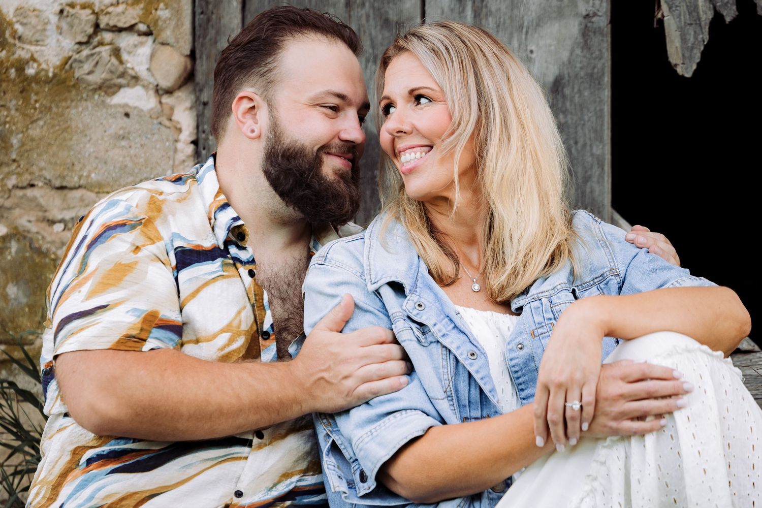 A smiling couple embraces outdoors, woman in denim jacket and white dress, man in floral shirt, rustic barn backdrop.