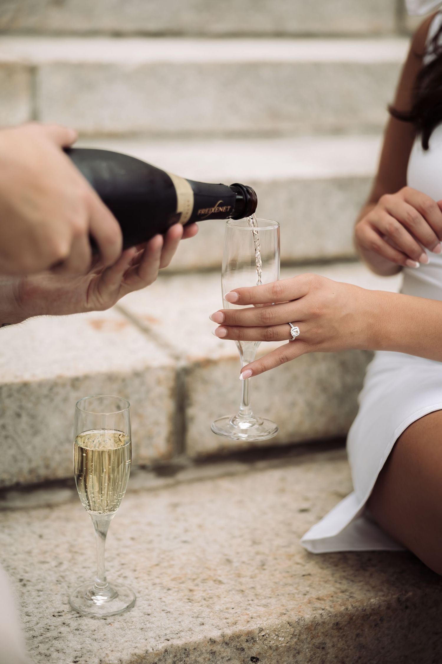 Champagne being poured into a flute held by a bride in a white dress, sitting on stone steps.