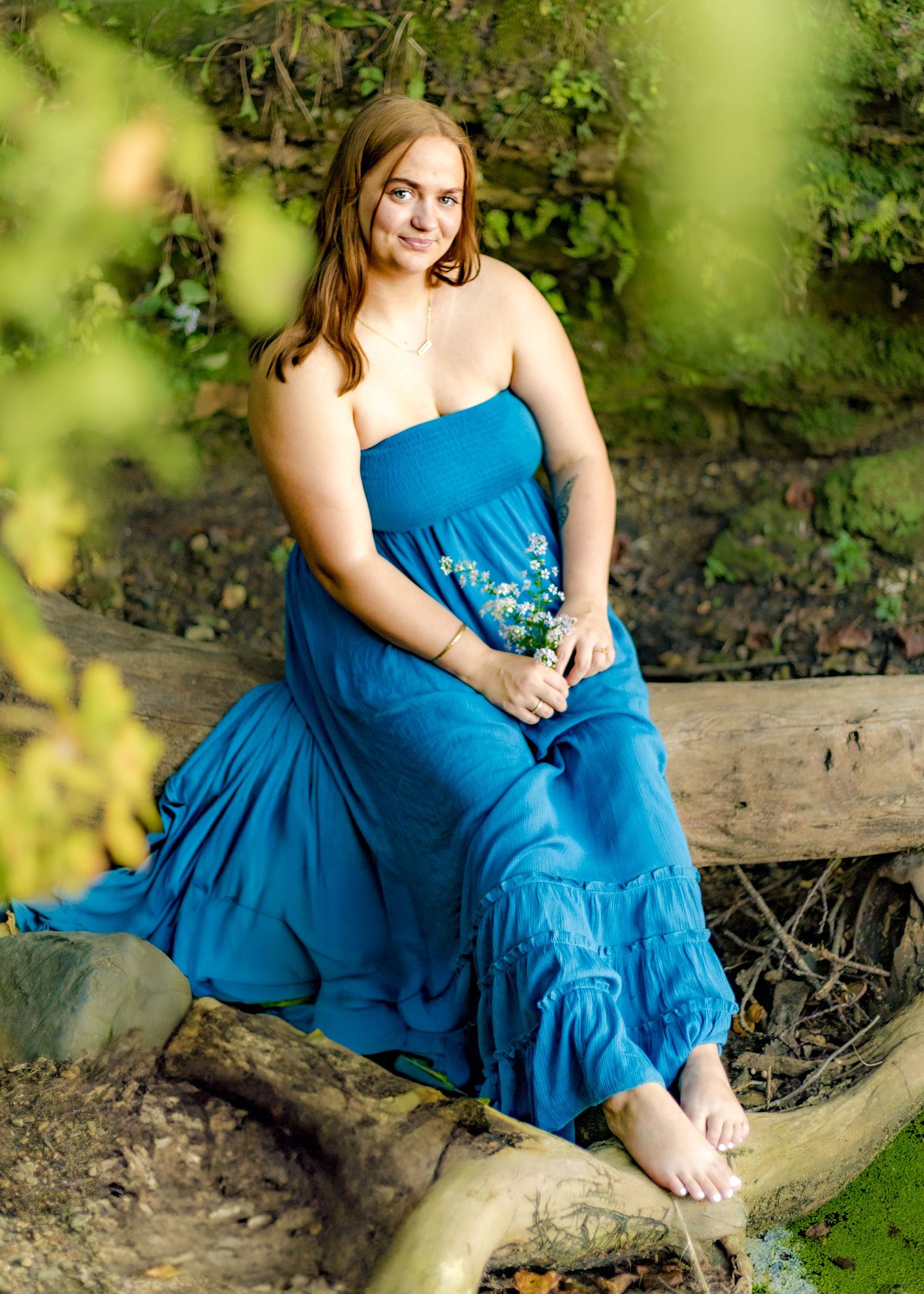 Young woman in blue strapless maxi dress sitting on log outdoors, holding wildflowers in a lush green garden setting.