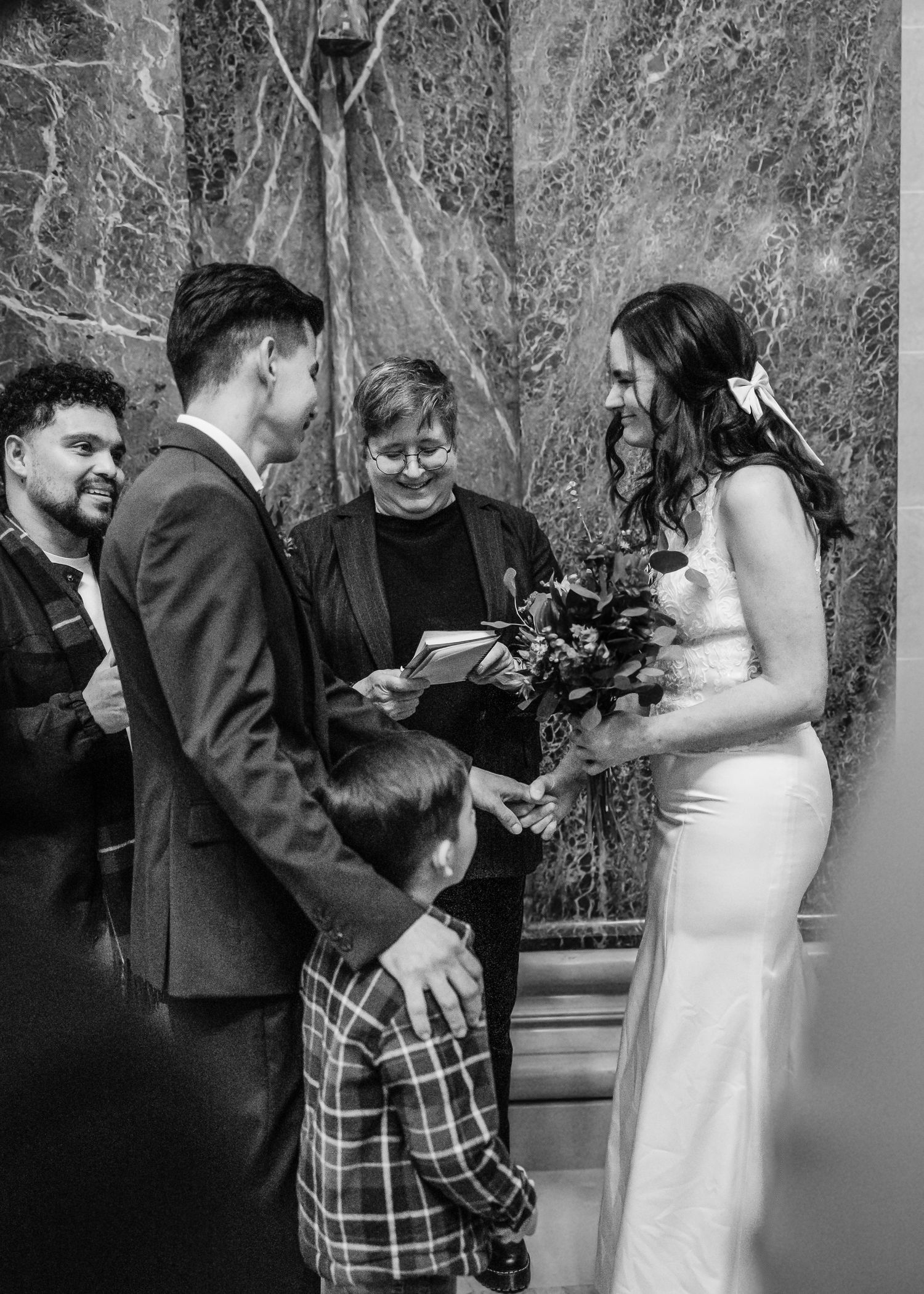 Bride and groom exchanging vows with officiant and child present at elegant marble venue.