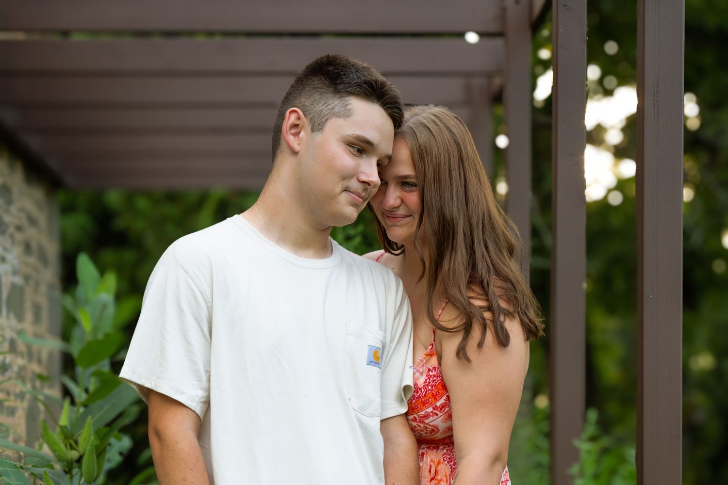 Romantic couple touching foreheads tenderly while standing together under a wooden pergola outdoors.