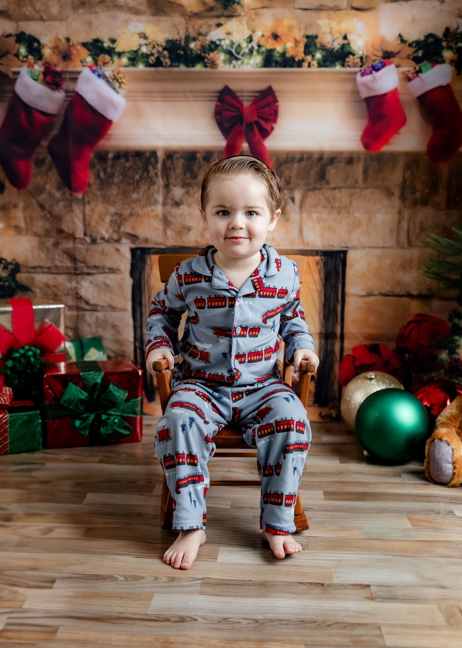 Toddler in blue train pajamas sitting in wooden chair by Christmas fireplace with stockings and gifts.