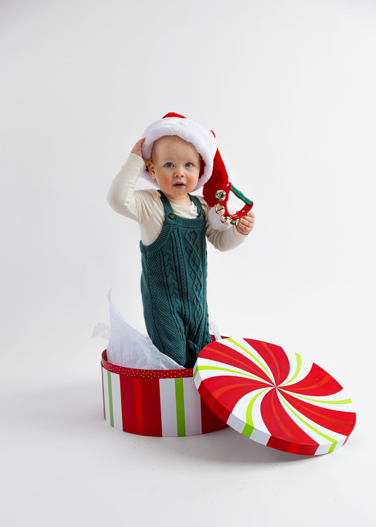 Curious baby in oversized Santa hat standing in large Christmas gift box holding holiday ornament.