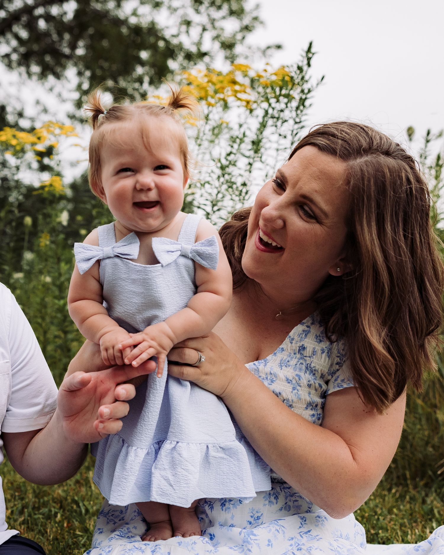 A joyful mother in a floral dress holds her laughing baby girl in a blue dress outdoors among wildflowers.