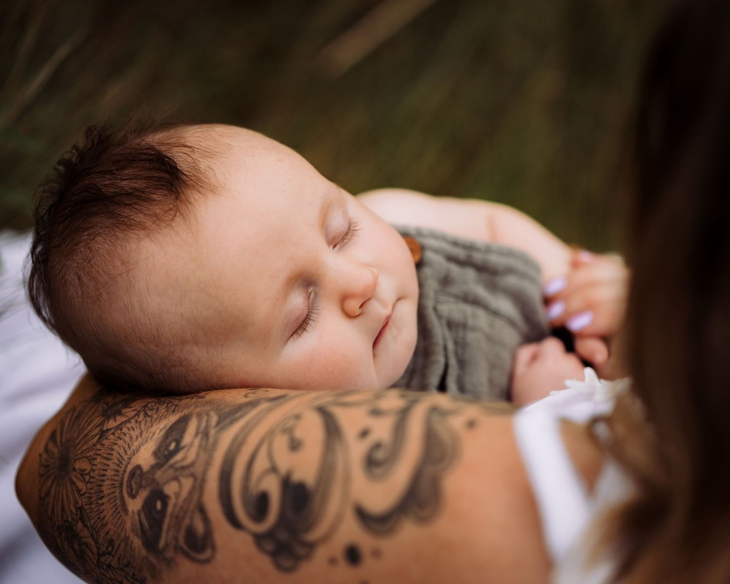 Sleeping newborn baby cradled on tattooed arm outdoors in natural setting.