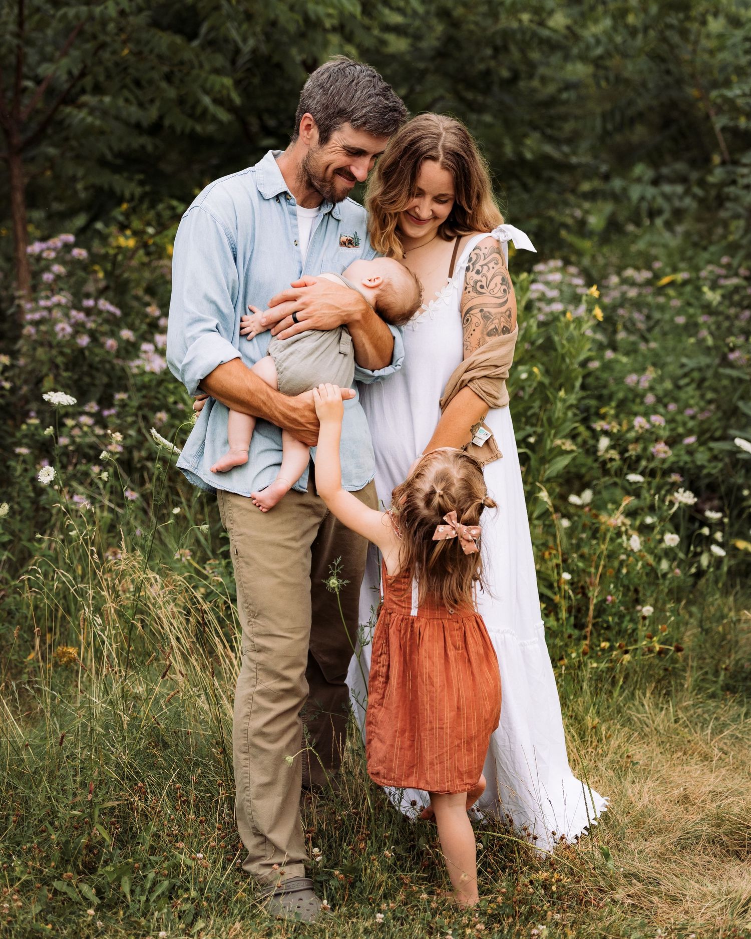 A happy young family with a newborn baby and toddler girl poses in a lush green meadow with wildflowers.