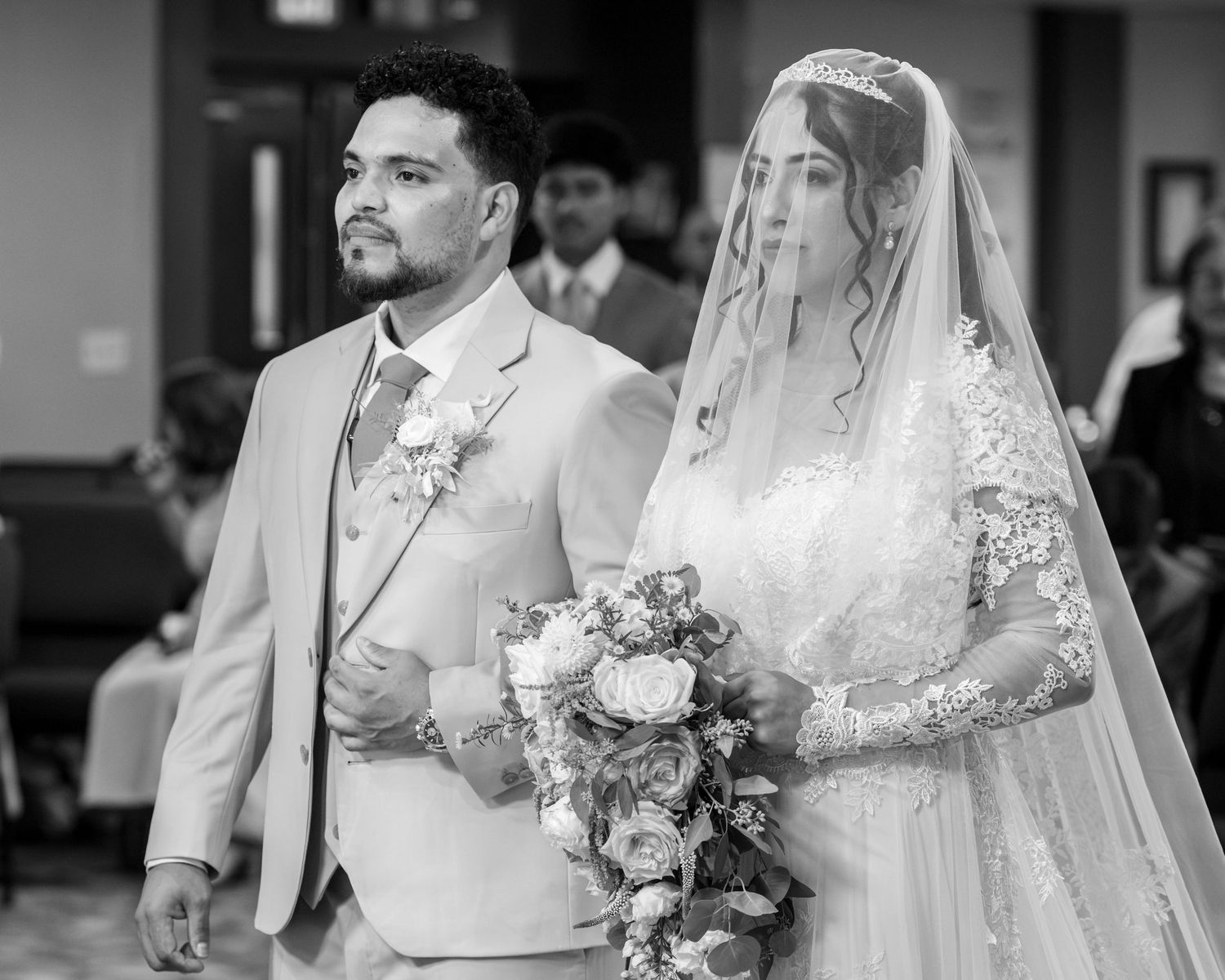 Black and white photo of a bride in a lace gown and veil holding a rose bouquet as she walks down the aisle beside a groom in a light suit.