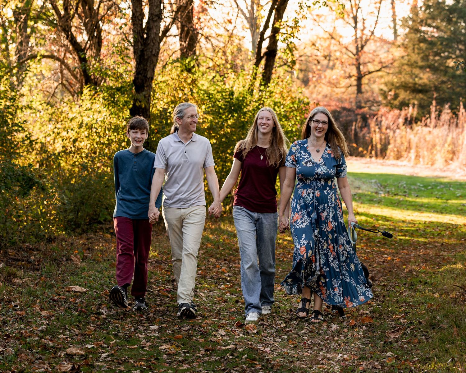Family of four holding hands and walking together along a leaf-covered path in a sunny autumn park surrounded by colorful trees.