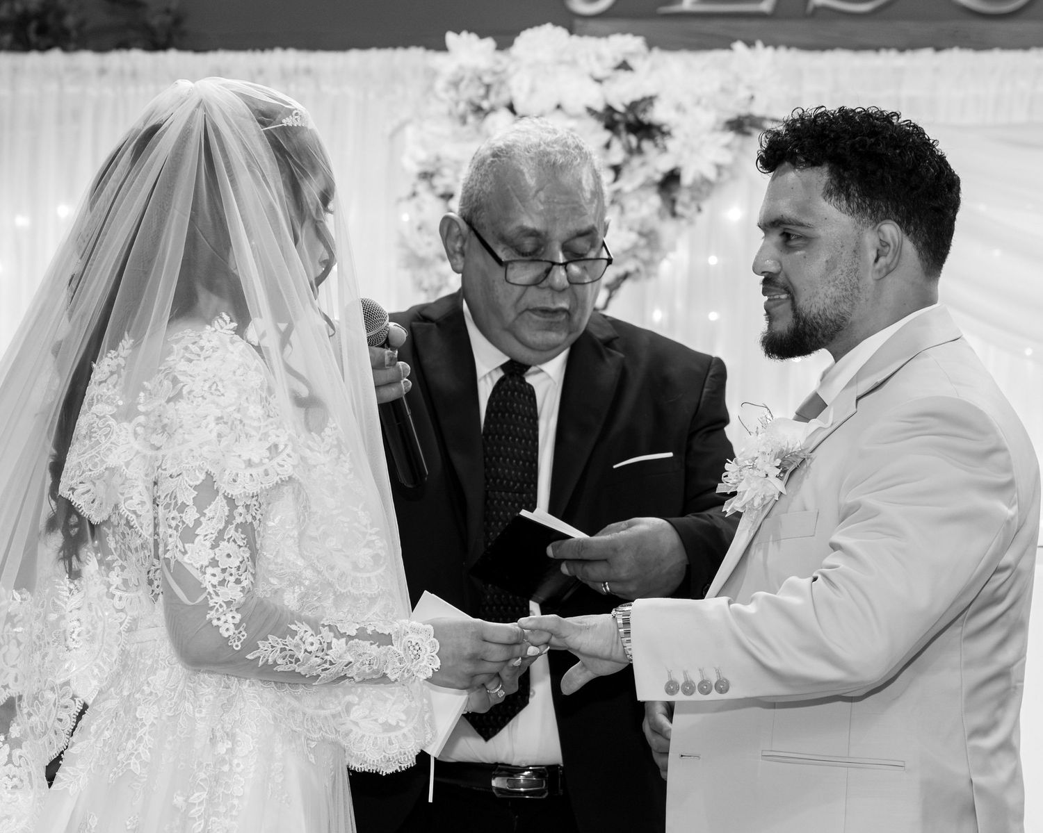 A bride in a lace gown and veil exchanges rings with a groom in a light suit as an officiant holds a book during their wedding ceremony.