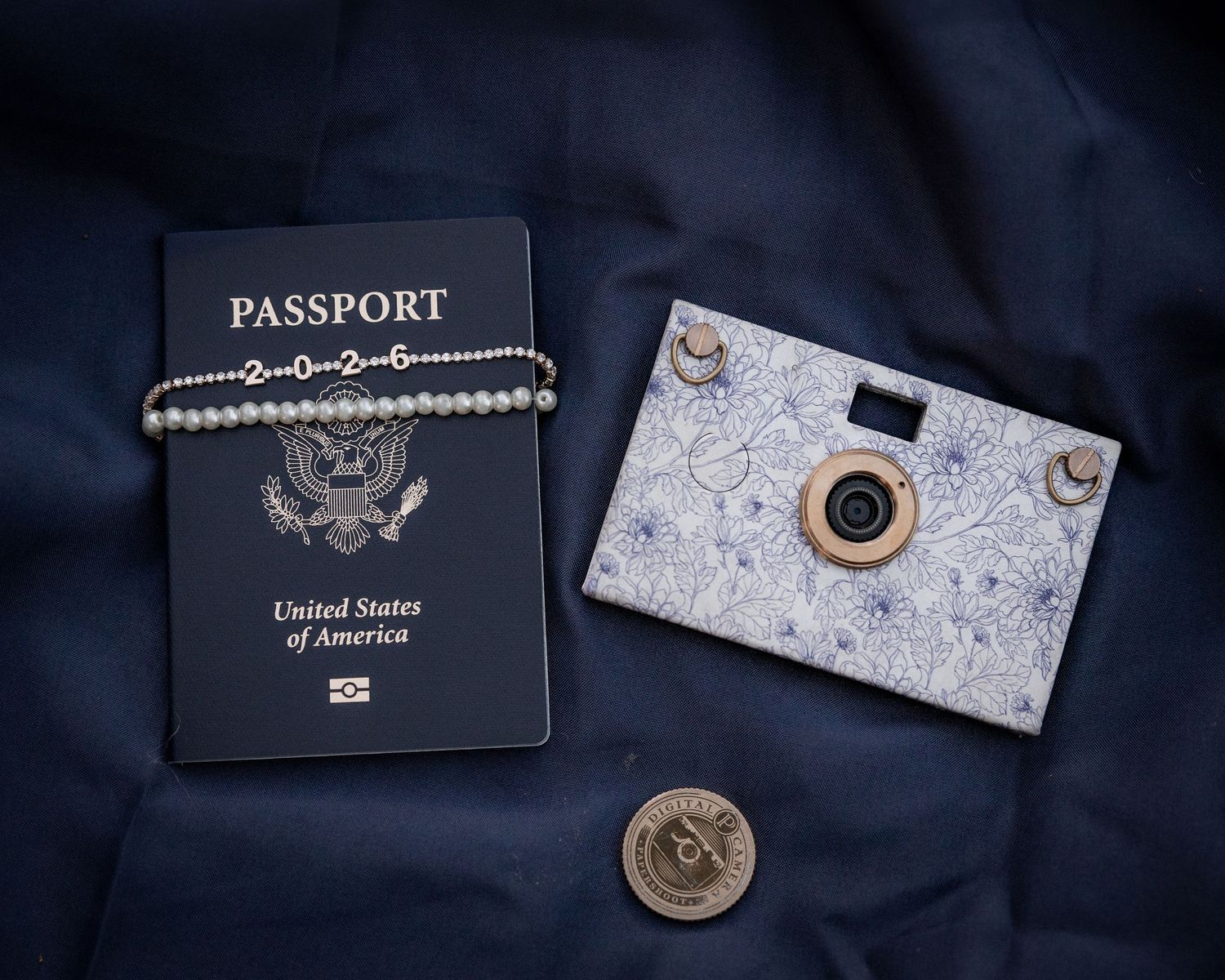 US passport, pearl bracelet, floral mini camera, and coin arranged on dark blue fabric for travel.