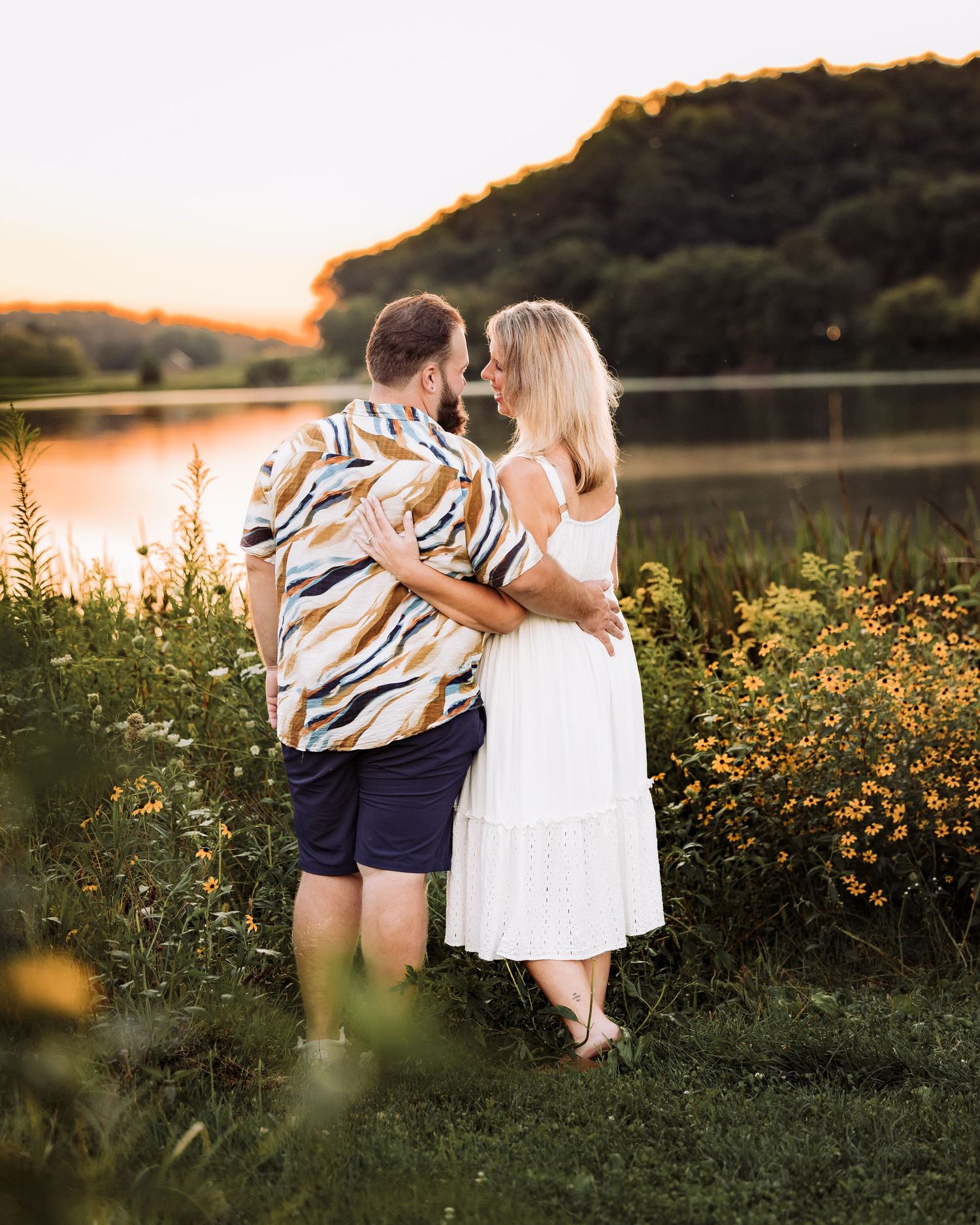 Couple embracing at golden hour by a serene lake, surrounded by wildflowers and lush greenery.
