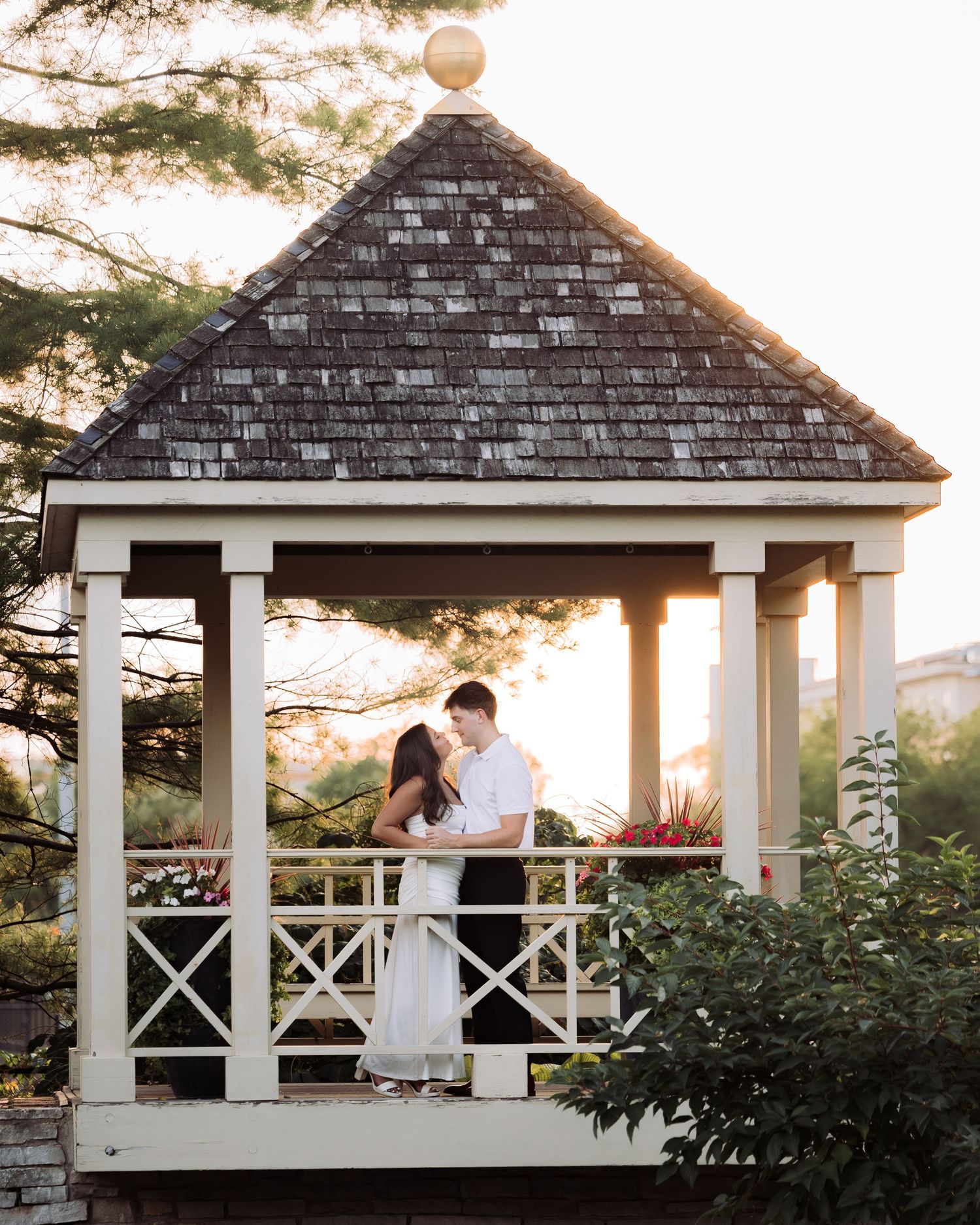 Couple sharing a romantic kiss inside a white gazebo with a shingled roof at golden hour sunset.