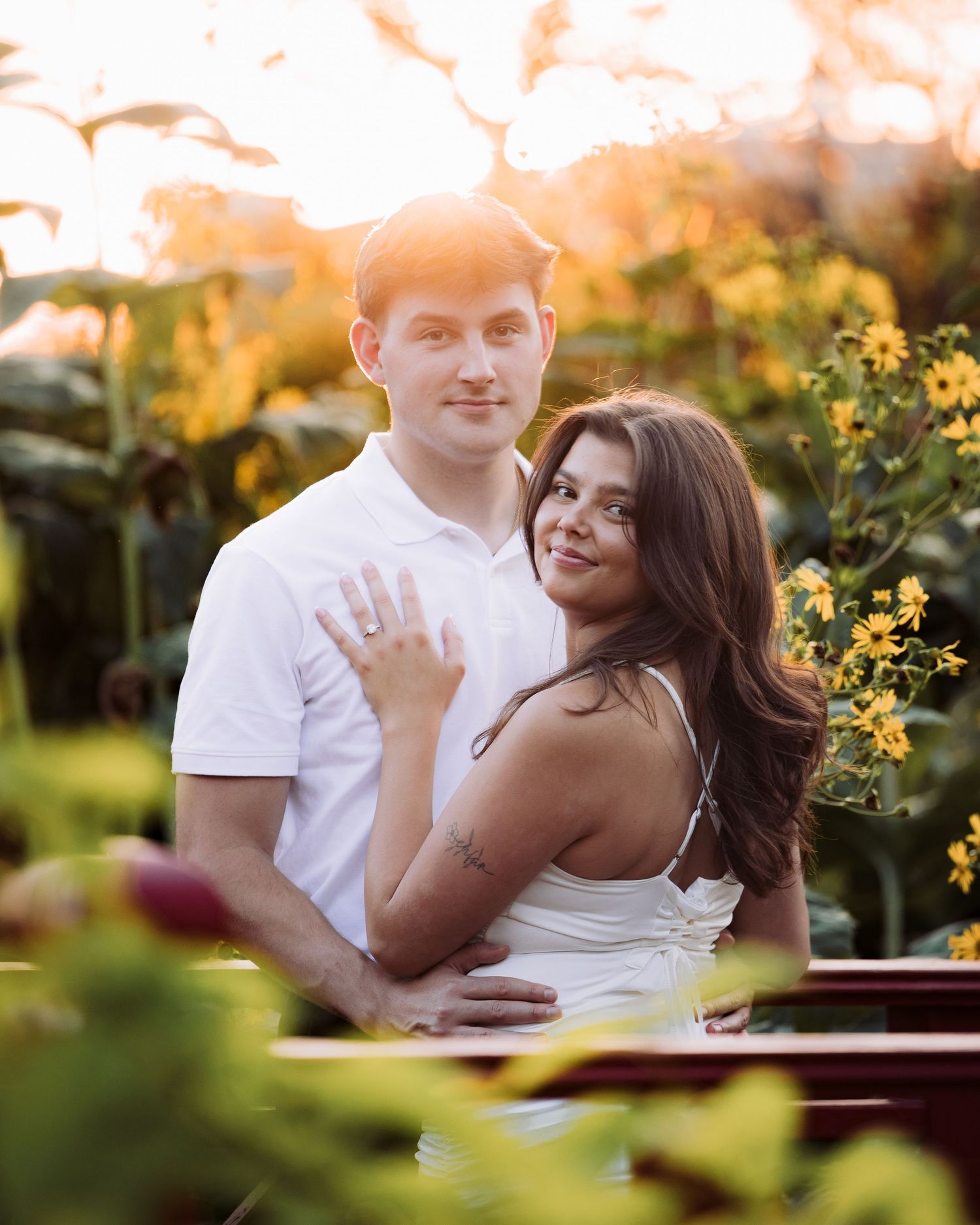 Couple posing at sunset in a garden, woman showing engagement ring, surrounded by yellow flowers and greenery.