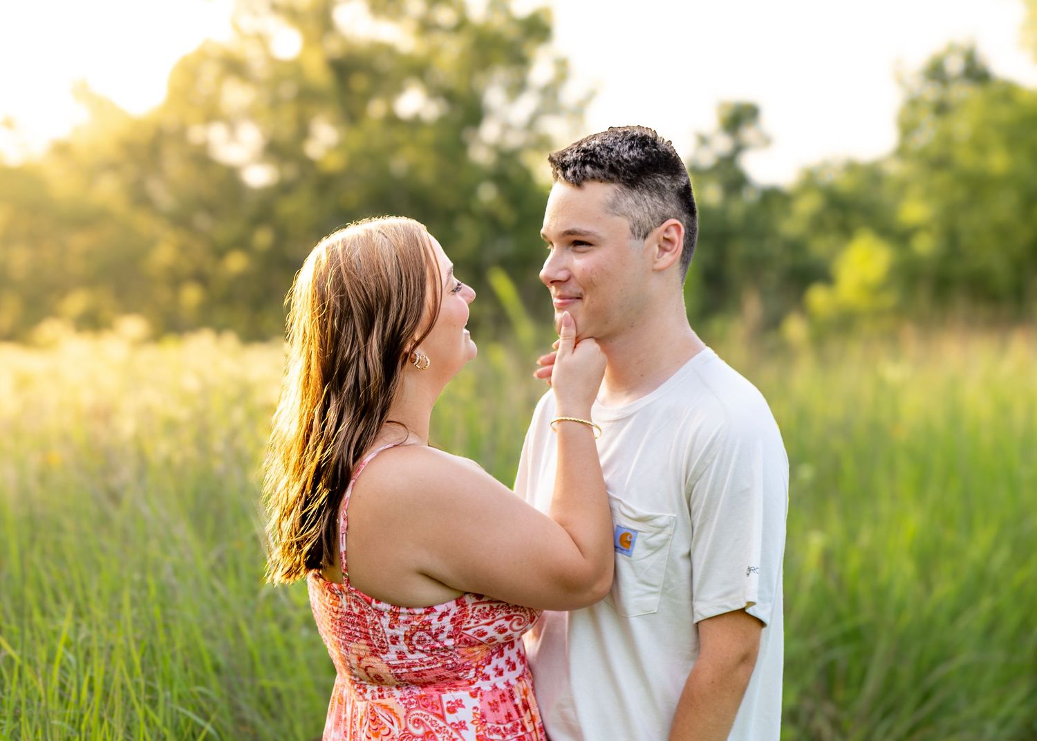 Couple gazing at each other and smiling in a sunlit green field, woman touching mans face gently.