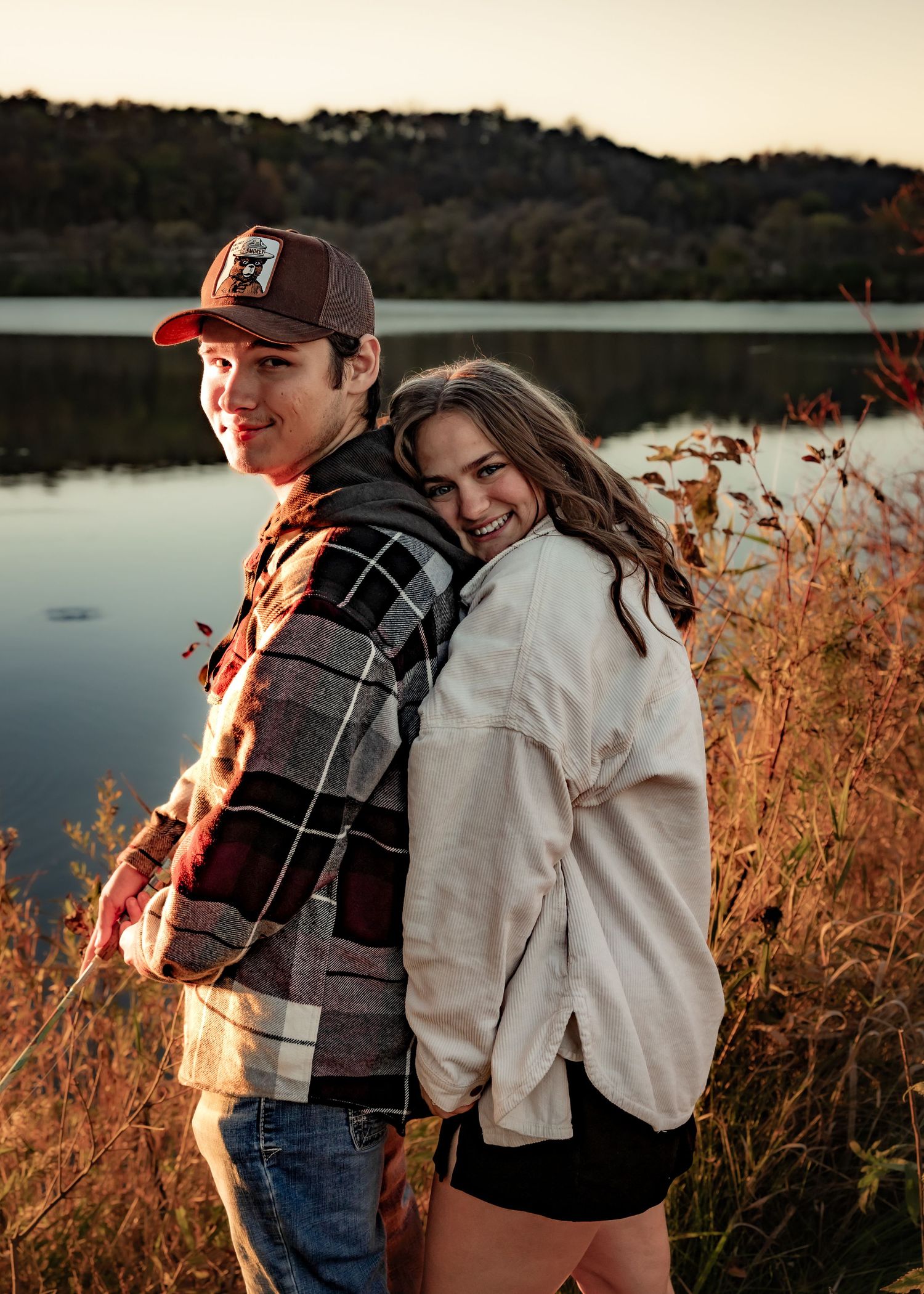 Smiling couple posing back-to-back near a lake with autumn fall foliage at sunset.