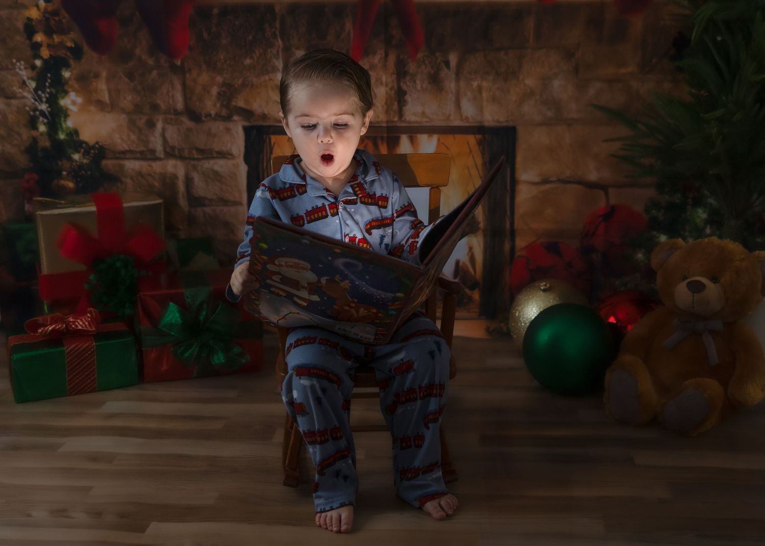 Young boy in train pajamas reading a glowing Christmas book by a decorated fireplace with gifts.