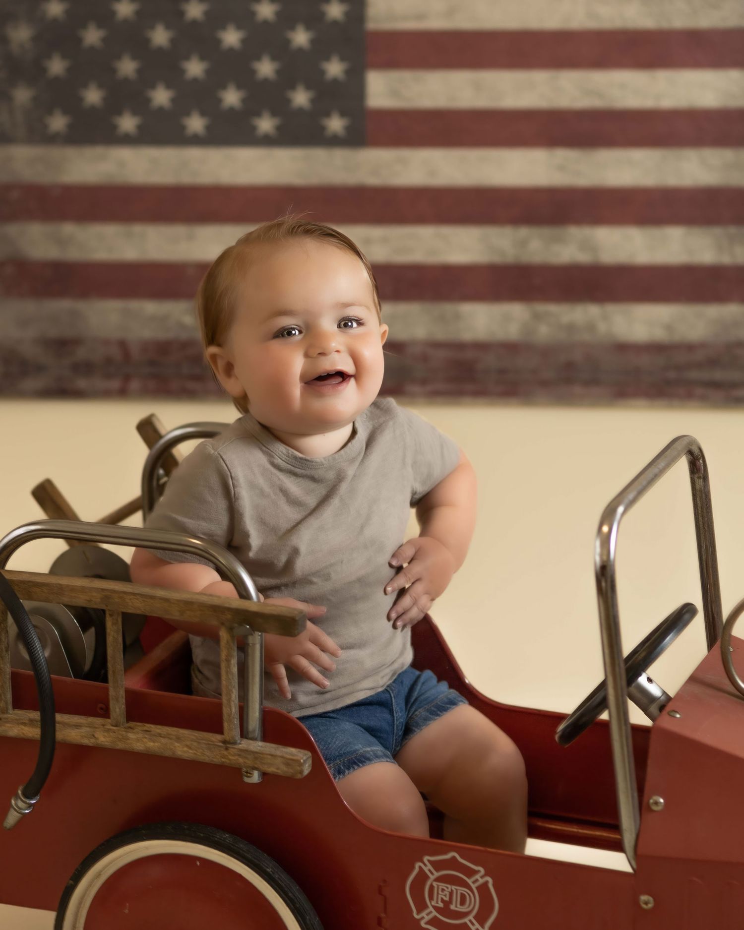 Happy baby in gray onesie sits in red vintage pedal fire truck with American flag backdrop.
