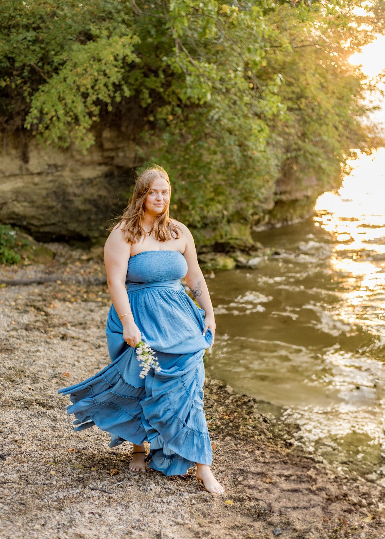 Young woman in blue strapless maxi dress walking barefoot along rocky shoreline at golden hour.