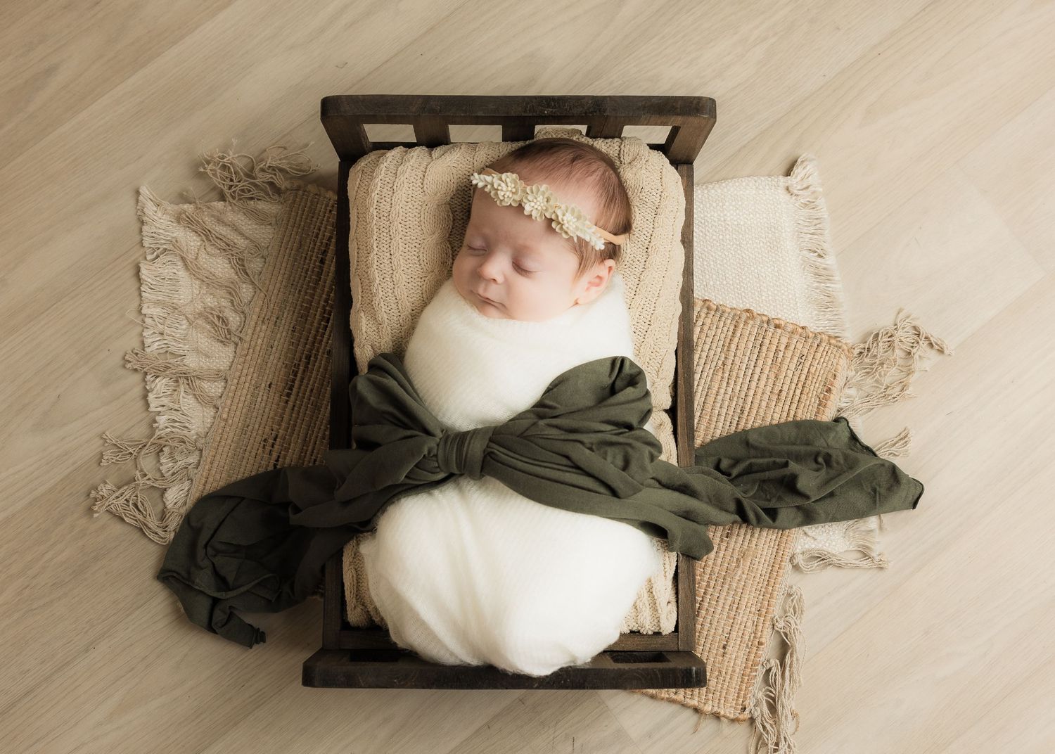 A sleeping newborn baby wrapped in white with a floral headband rests in a rustic wooden mini bed.