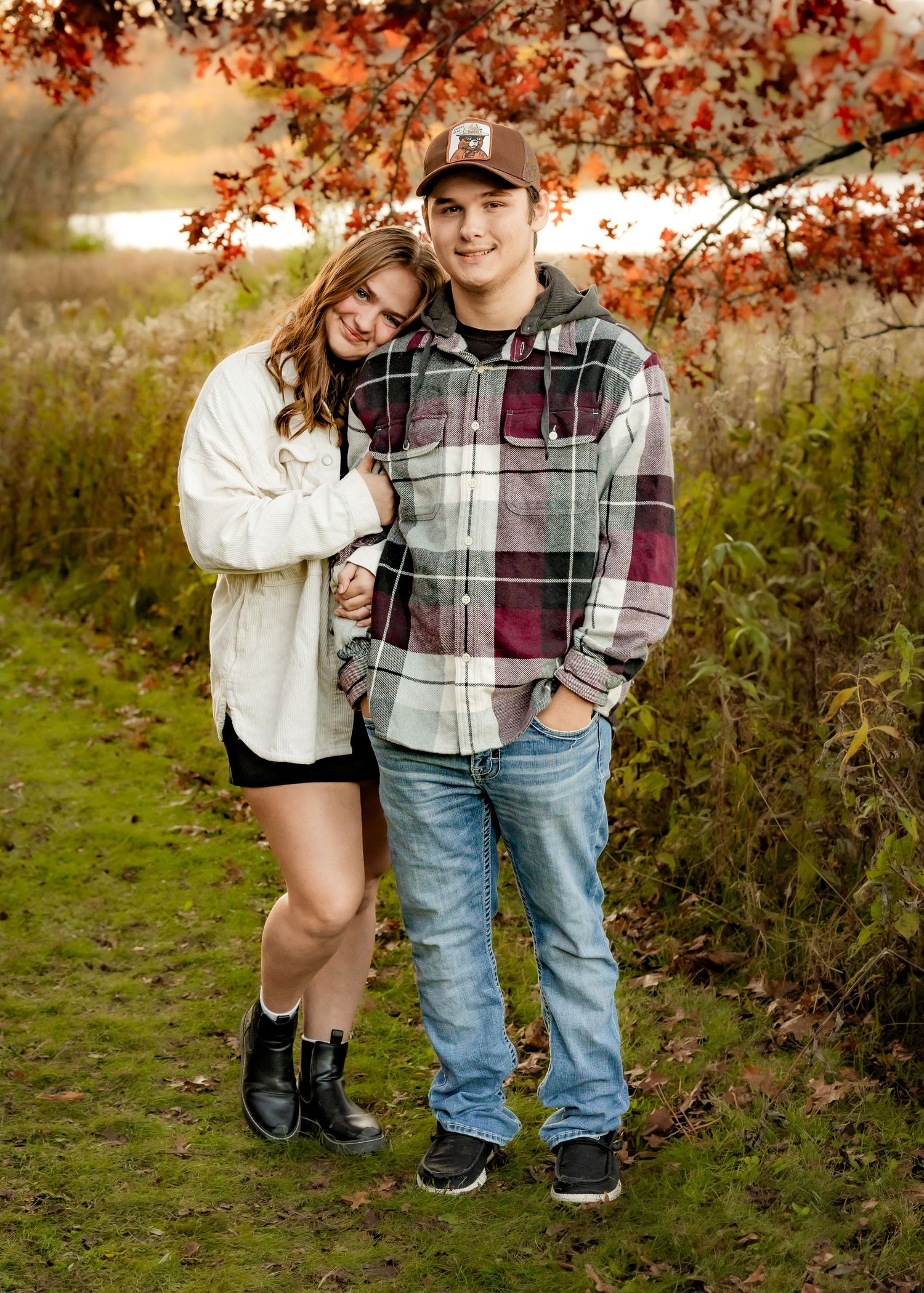 A young couple embraces outdoors beneath vibrant autumn foliage near a serene lake backdrop.