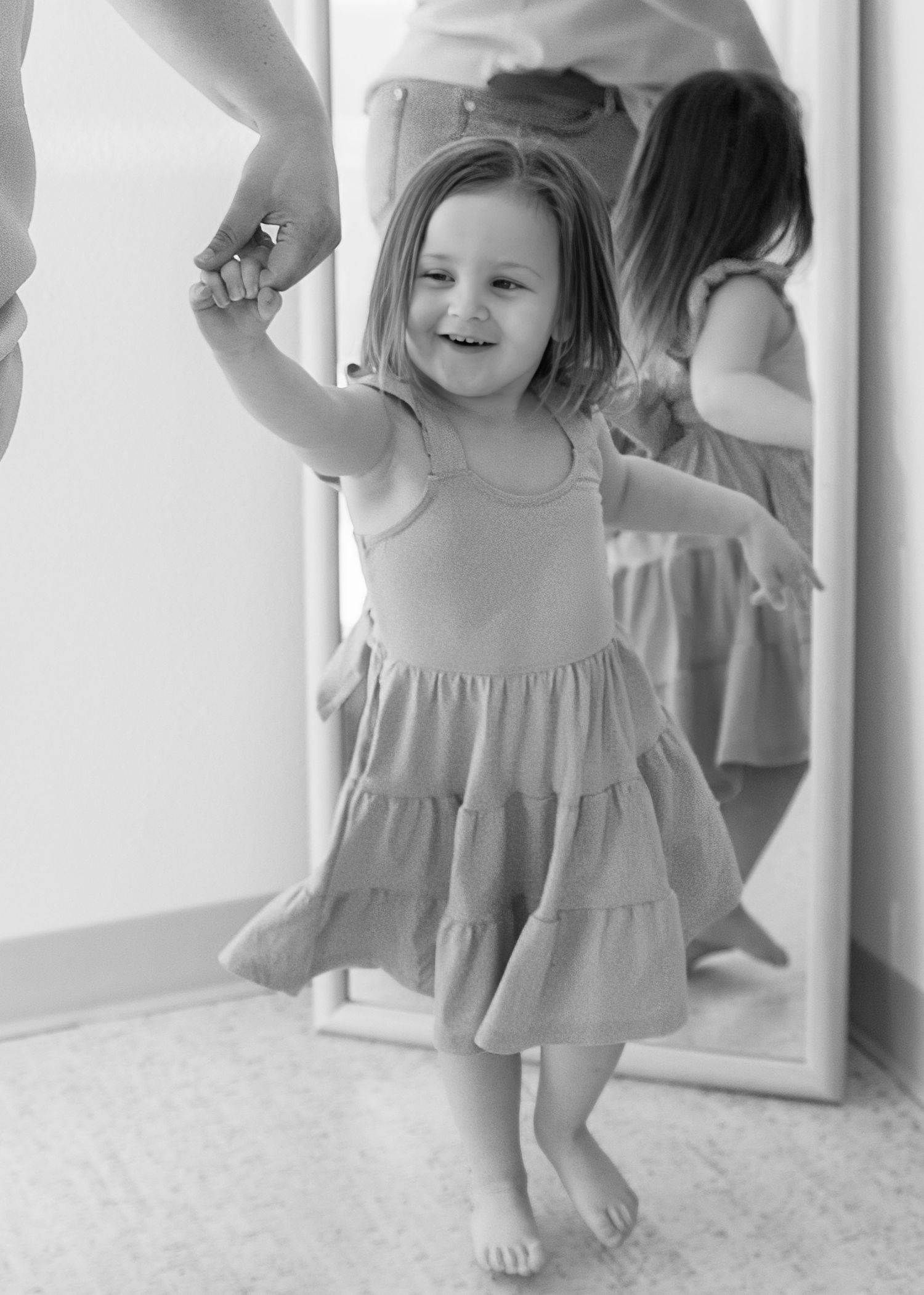 A joyful toddler girl in a tiered dress dances barefoot near a mirror in a black and white photo.